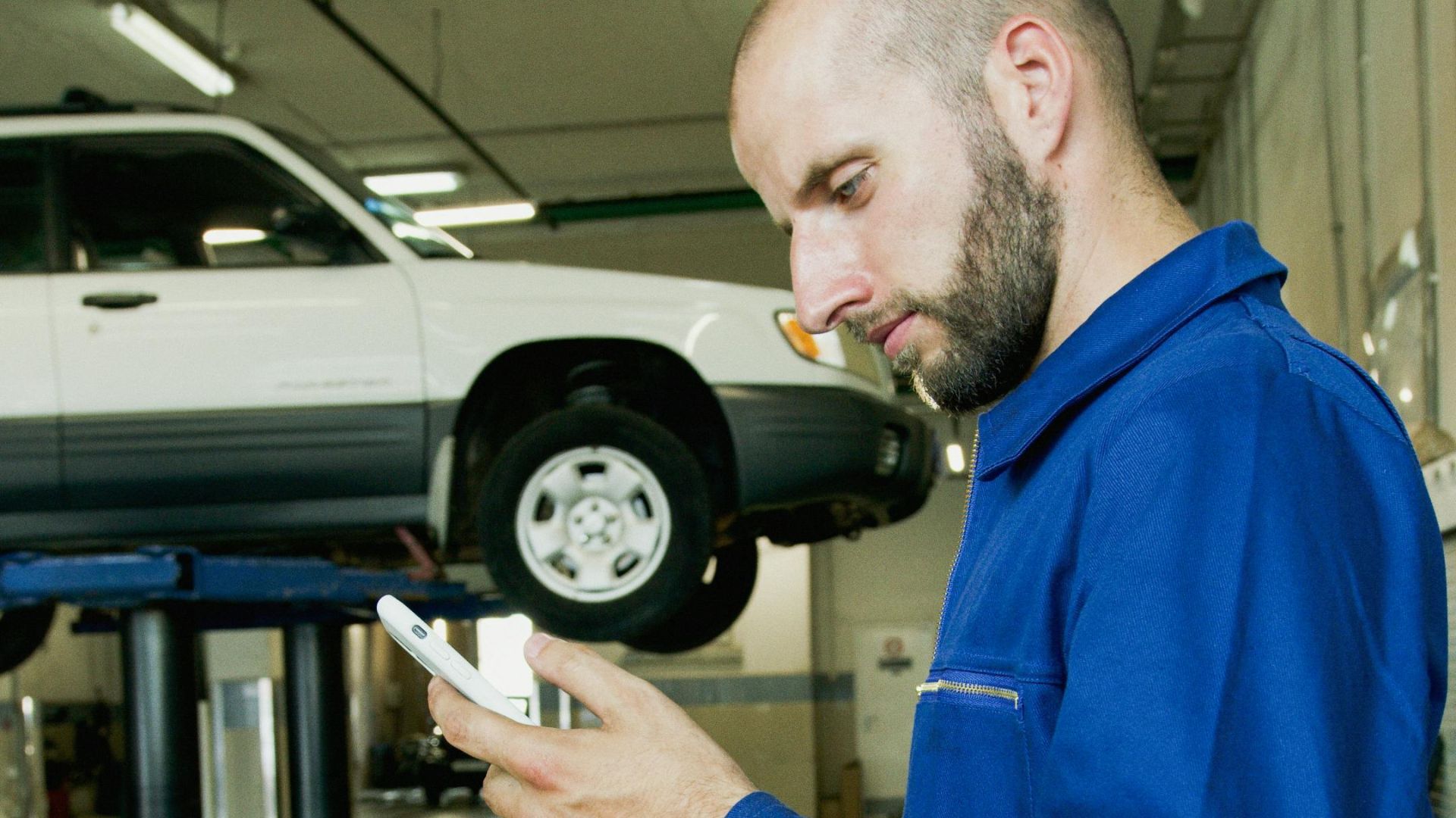 Bald mechanic in blue jumpsuit using phone in an auto repair garage with a car on the lift.
