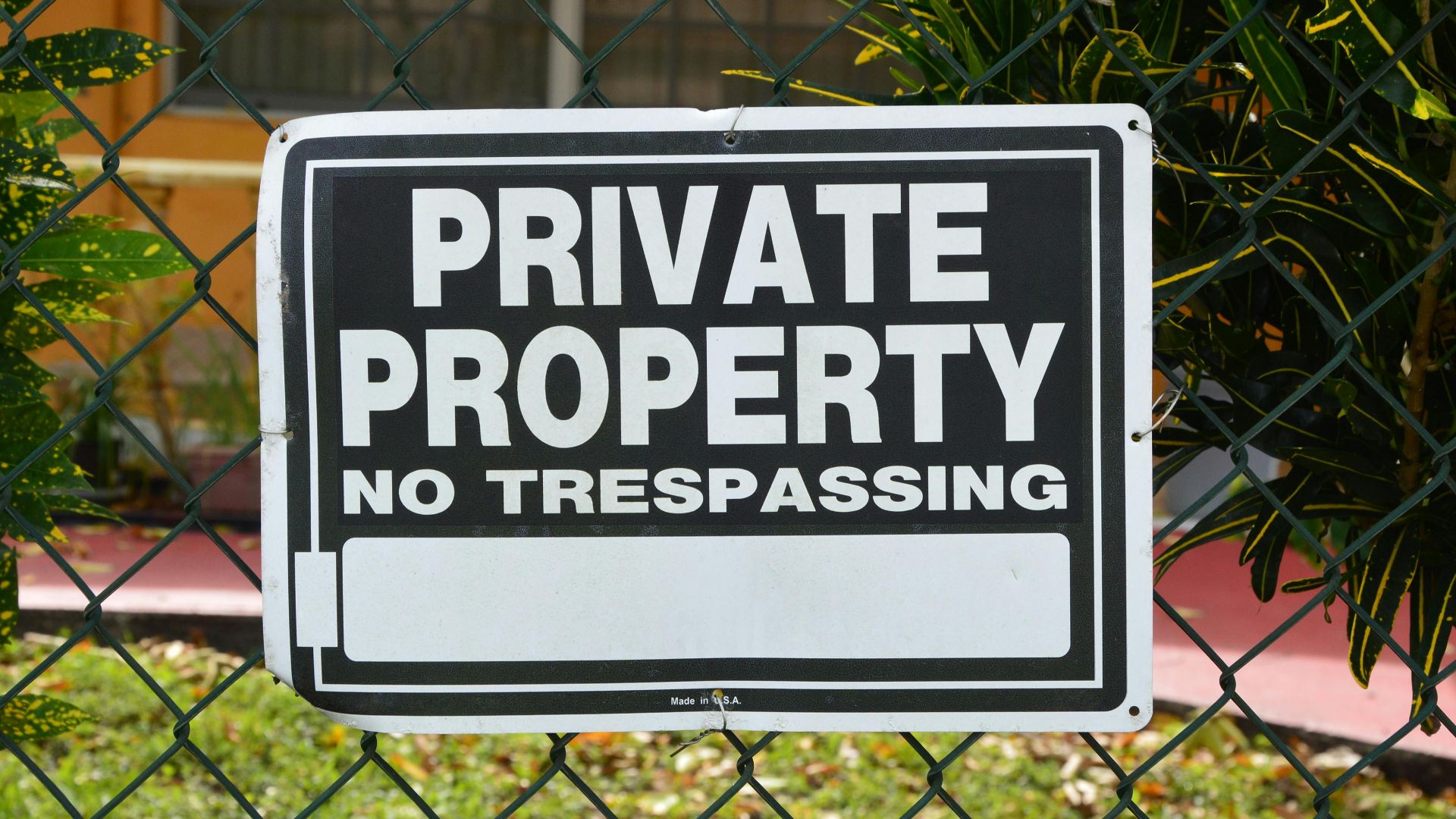 Close-up of a private property no trespassing sign on a chain-link fence with greenery in the background.
