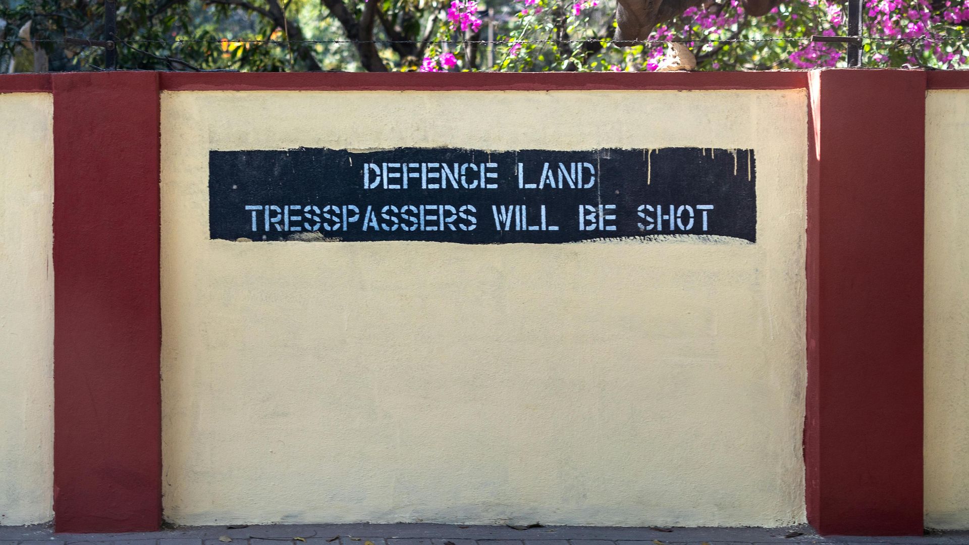 A warning sign reads 'Defence Land Trespassers Will Be Shot' on a wall in Bengaluru with blooming flowers.