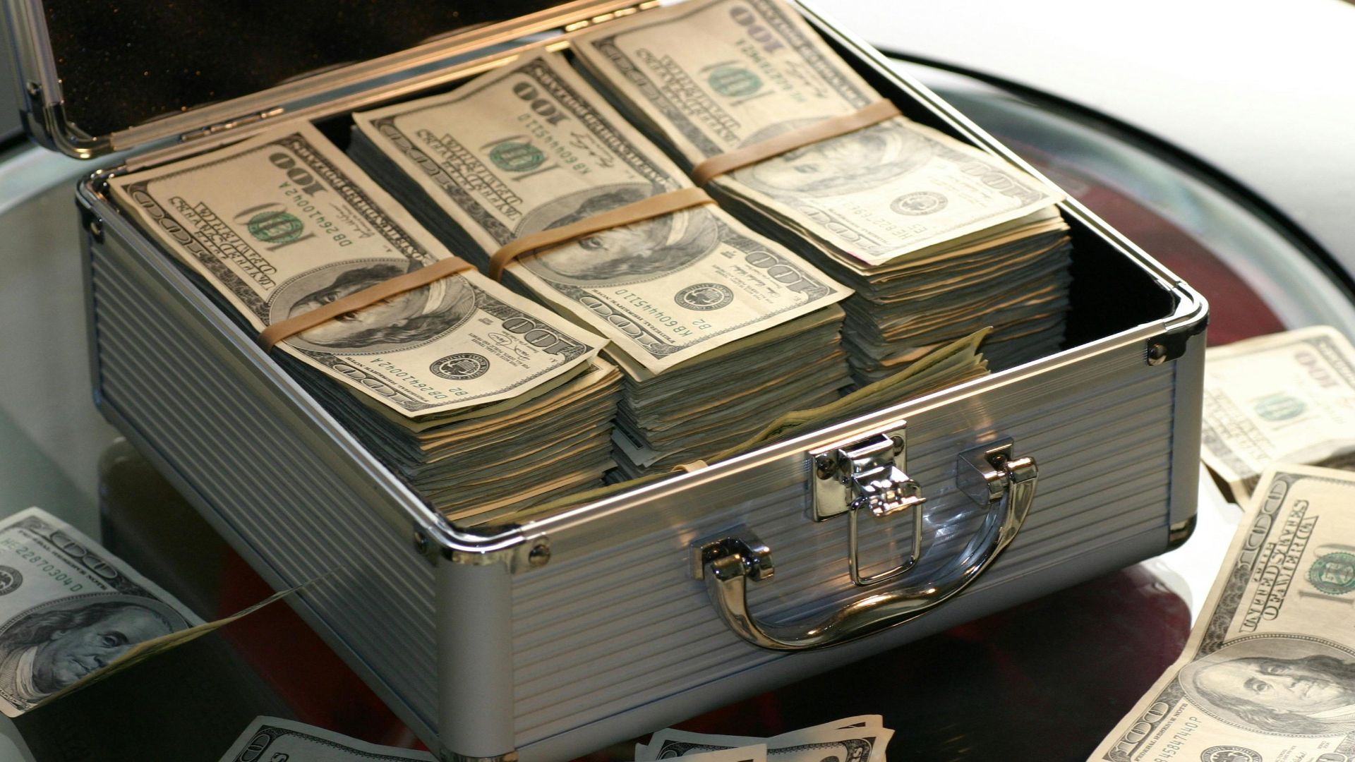 Open briefcase filled with stacks of hundred dollar bills on a glass table, representing wealth.