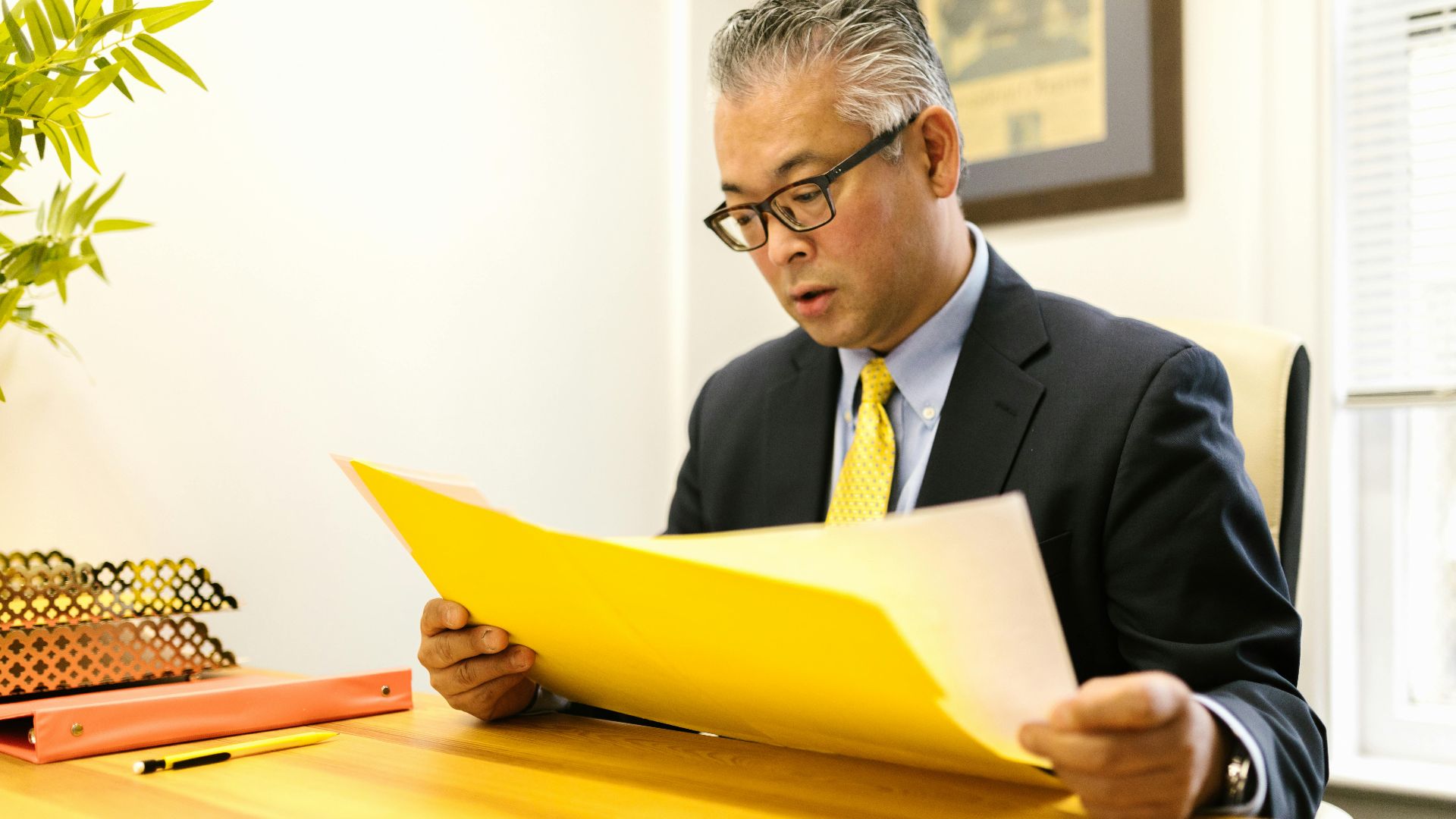 Asian businessman in corporate attire reading documents at office desk with a yellow folder.