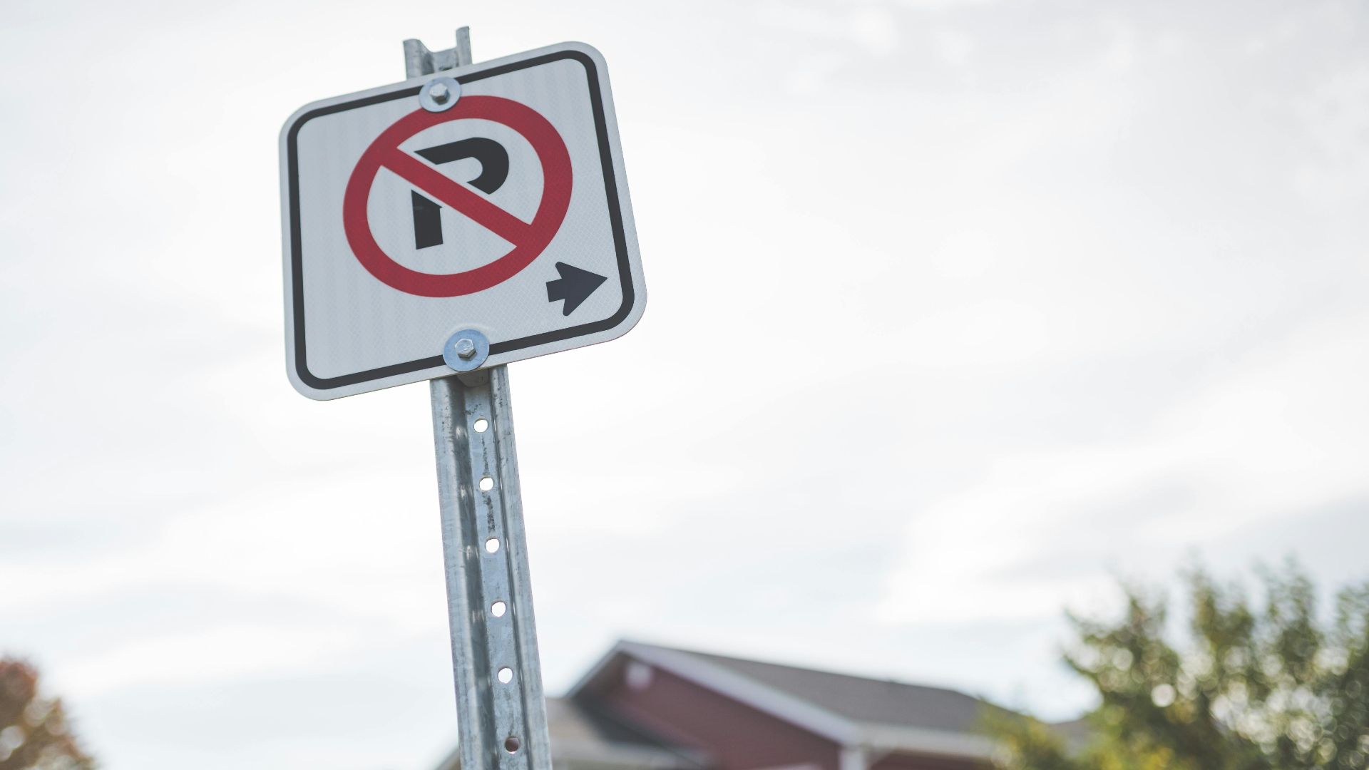 A no parking sign stands on a suburban street with a house and trees in the background.