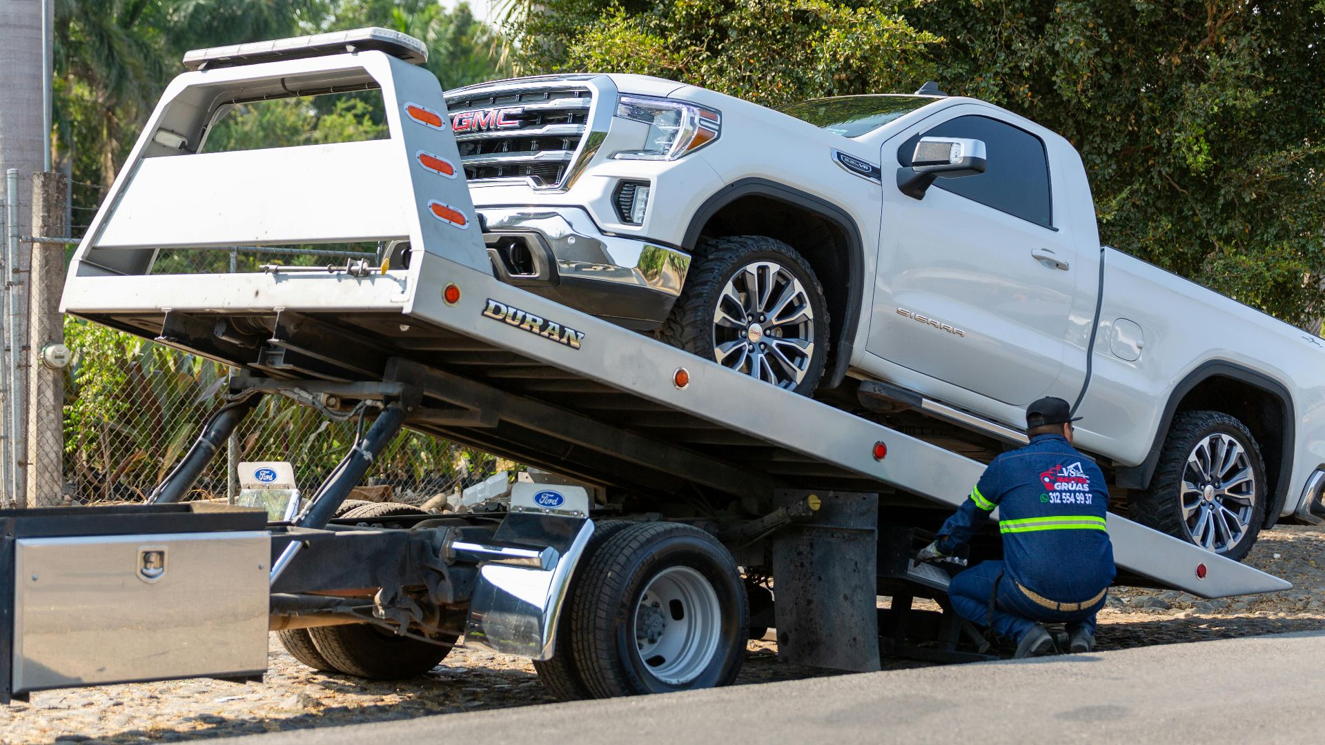 Tow truck operator loading white GMC pickup truck on street in daytime.