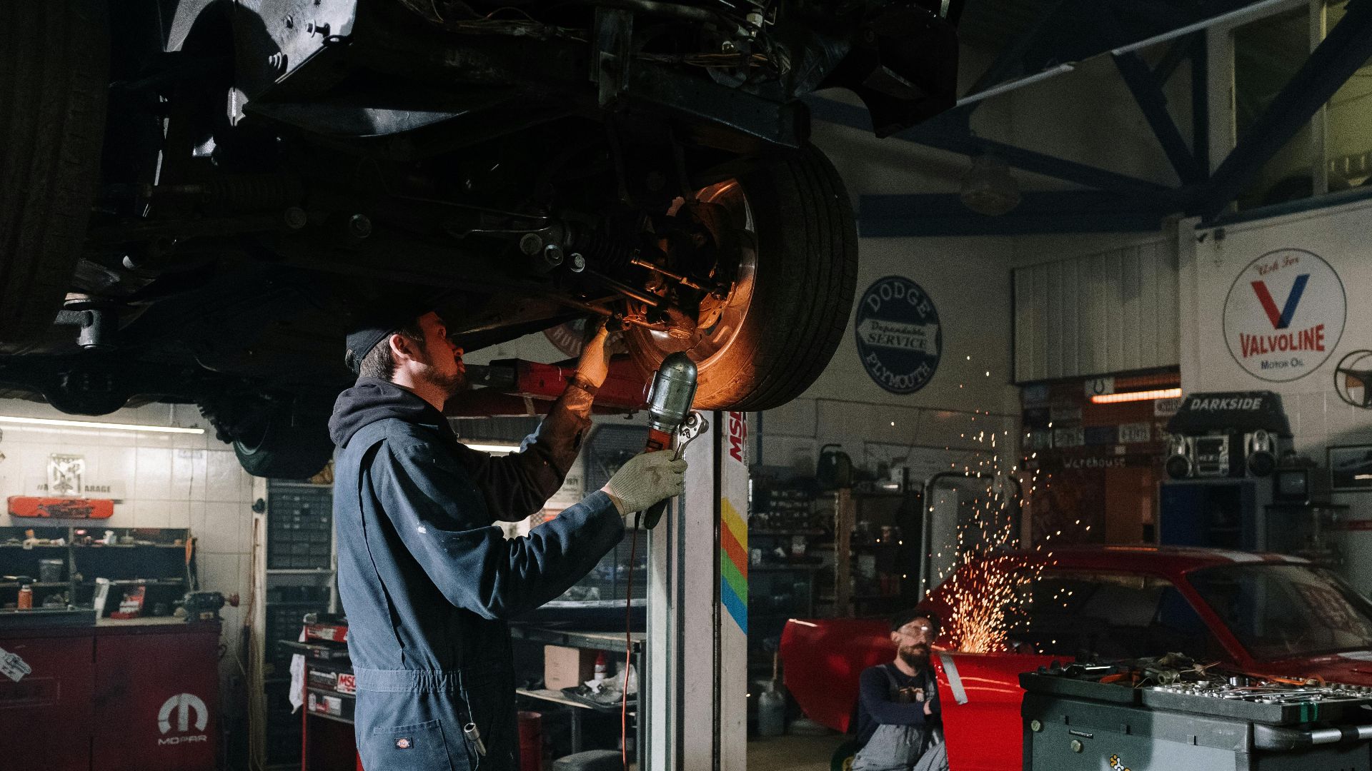 Two automotive technicians repairing a car in a busy workshop with sparks flying.