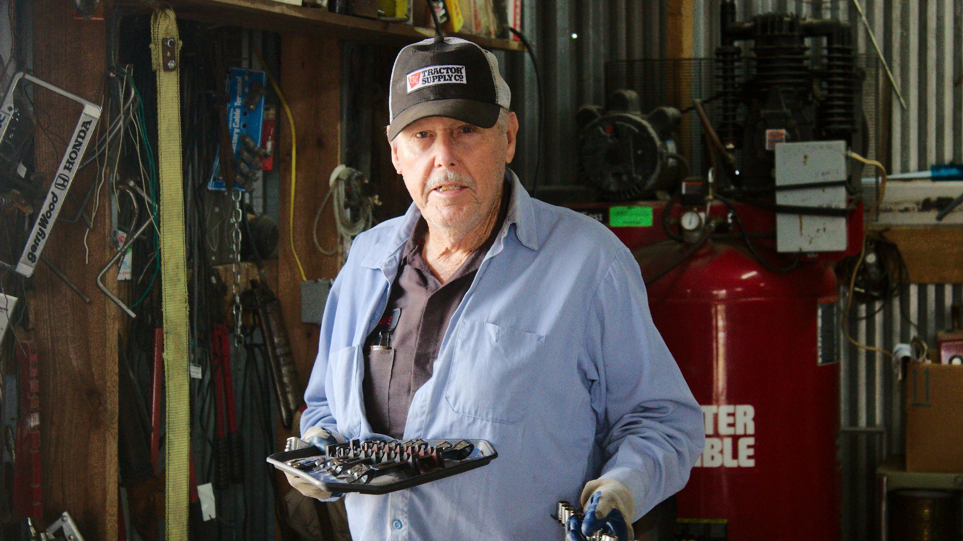 Portrait of an older mechanic holding tools inside a workshop setting, surrounded by equipment.