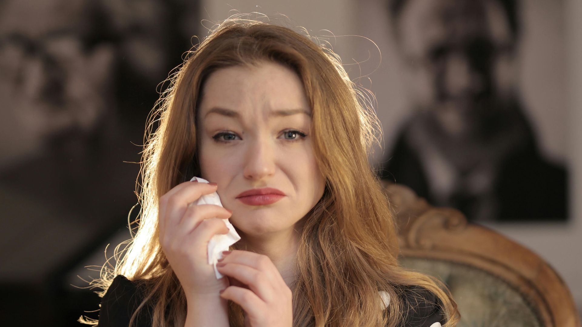 A woman sitting in a vintage chair indoors, showing emotions and holding tissues.