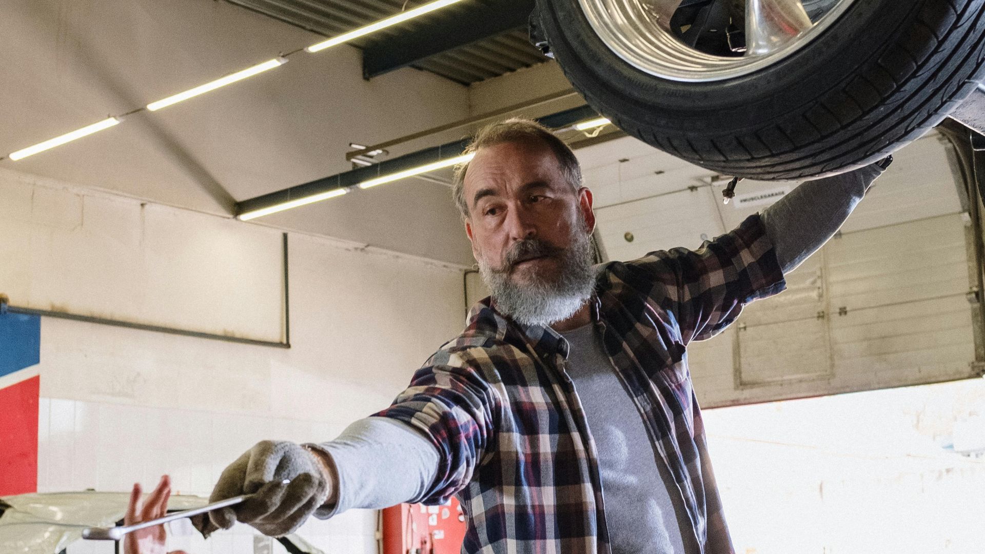 Bearded mechanic working on a vehicle in an auto repair shop, handing a tool to an assistant.