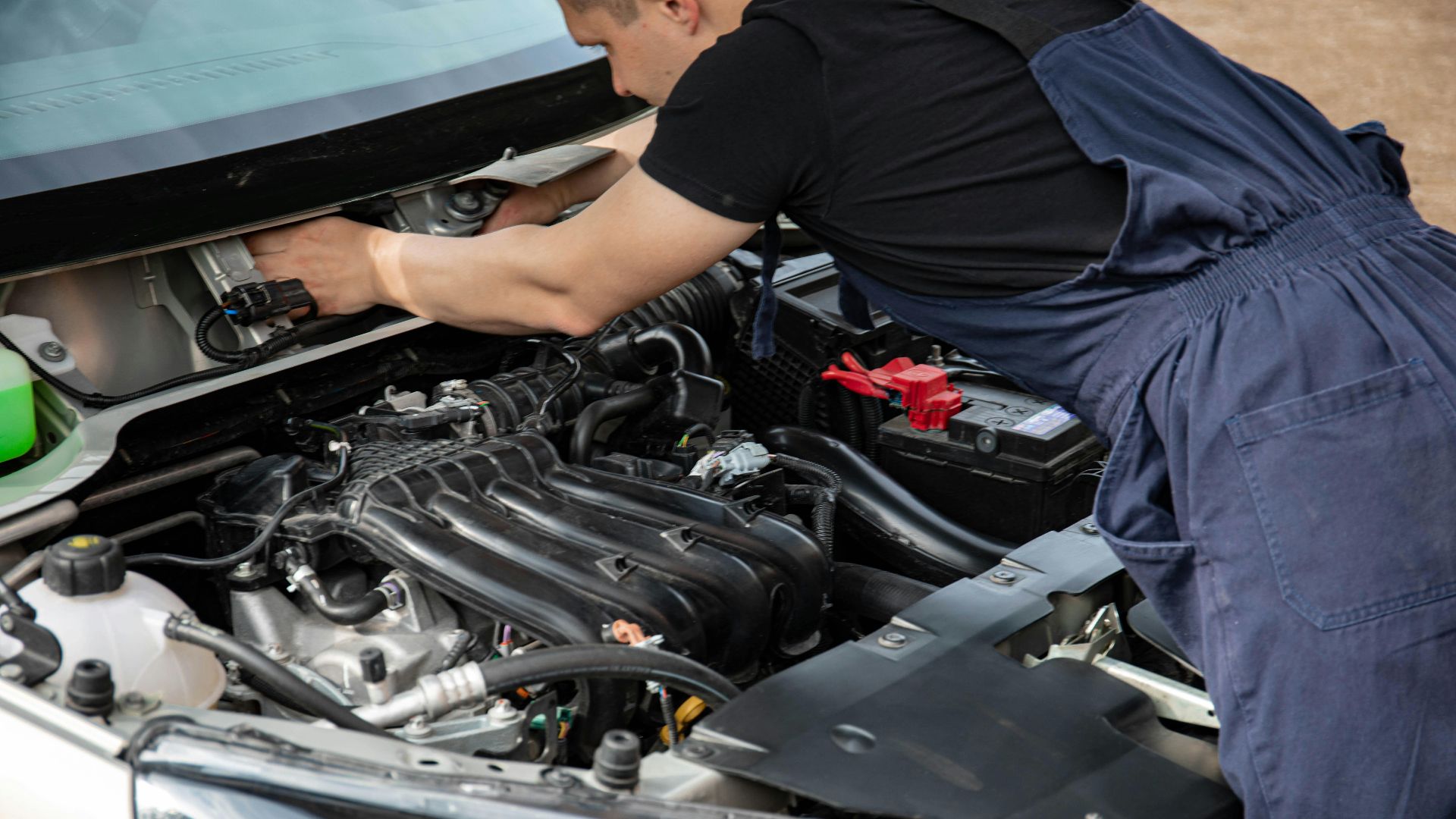 Mechanic in blue overalls working on car engine with open hood outdoors during the day.