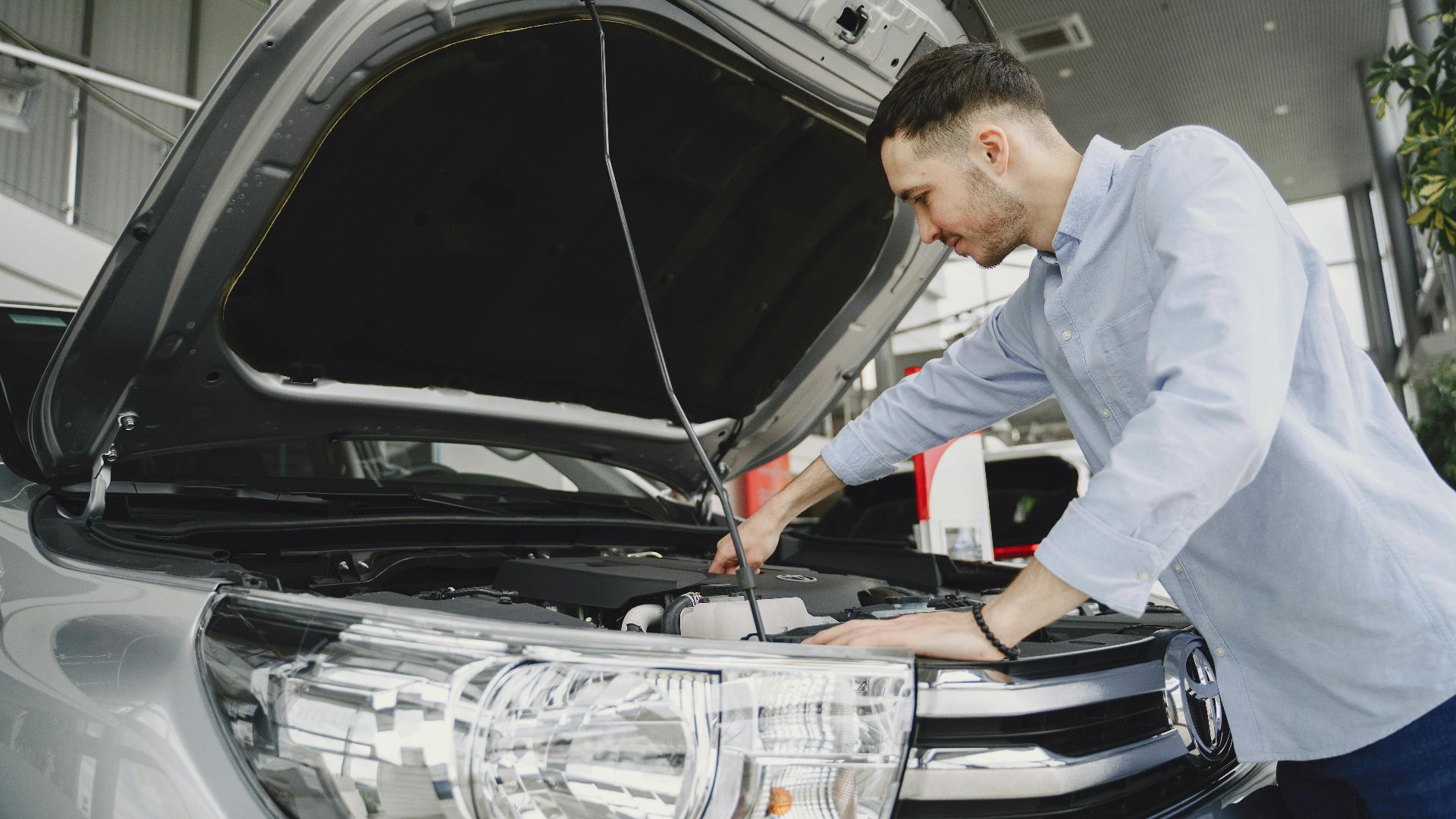 A man in a blue shirt examines a car's engine in a modern vehicle showroom.