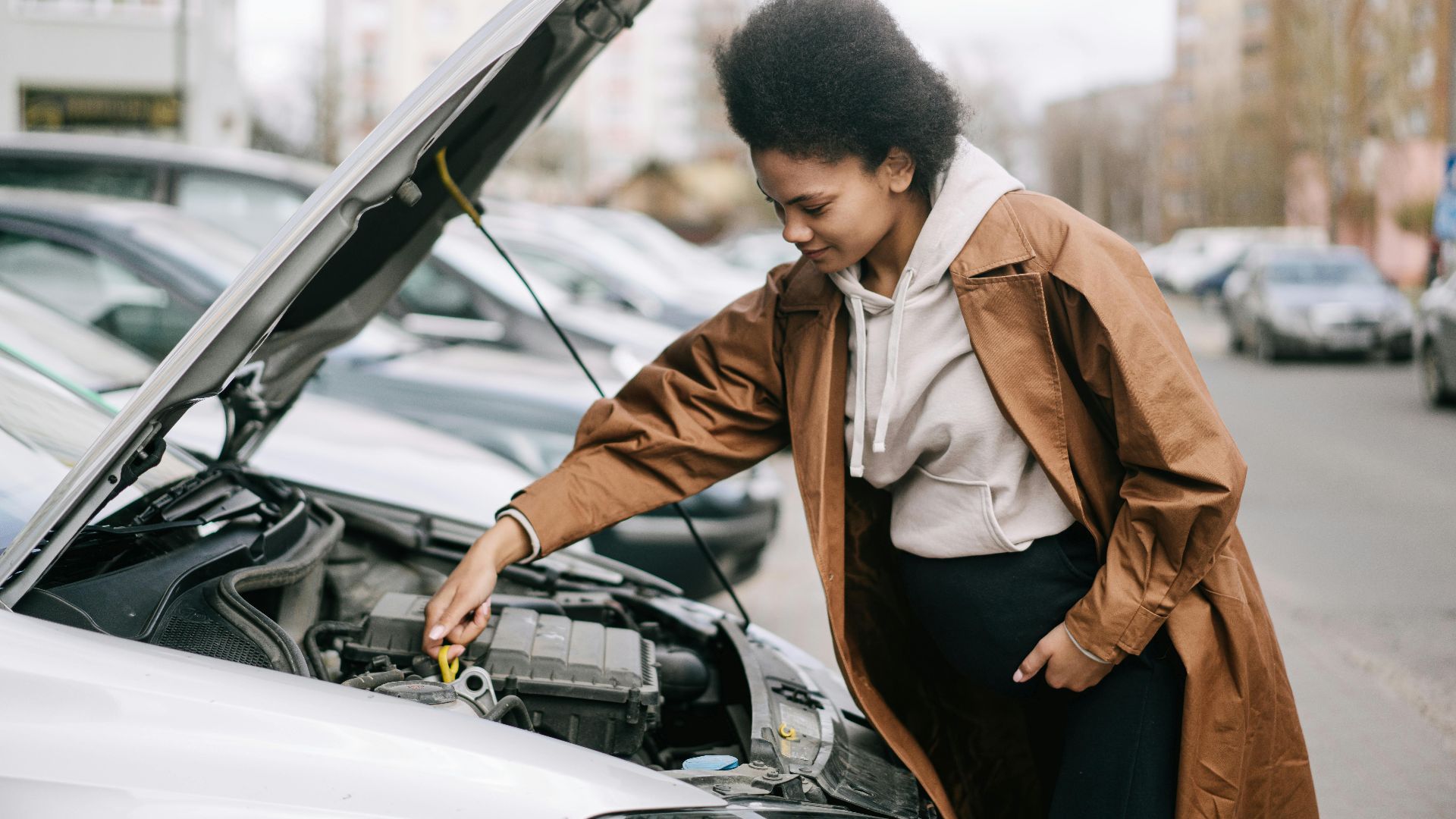 A pregnant woman inspects a car engine outdoors, showcasing independence and car maintenance skills.