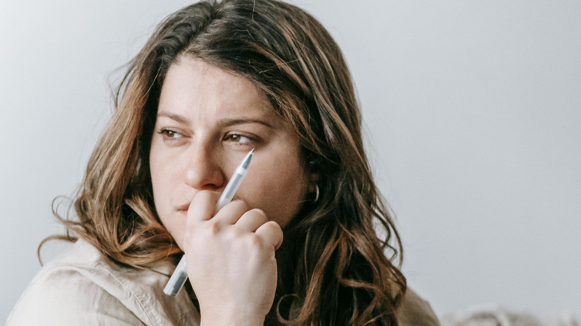 A woman holds a cup and pen, looking contemplatively indoors by a laptop.