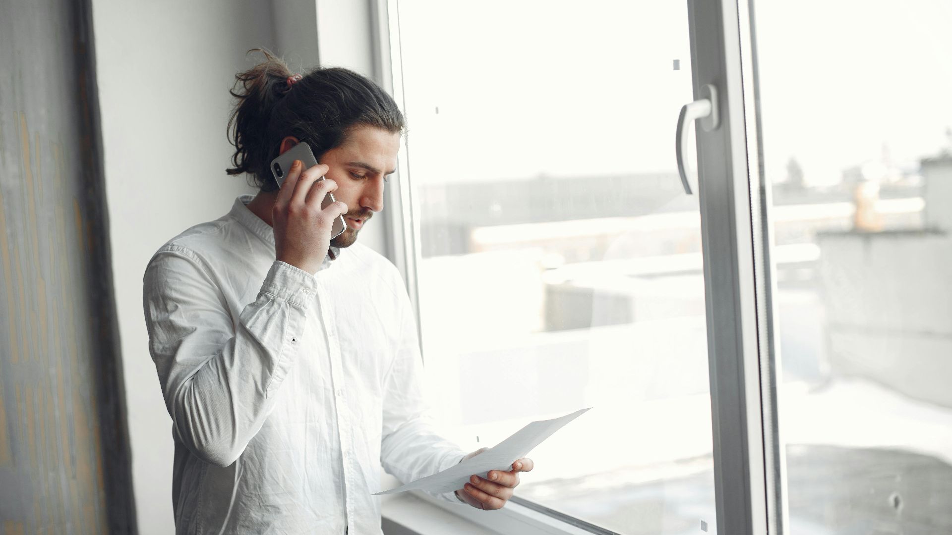 Young man in white shirt, on phone call holding a document, standing by a large window.