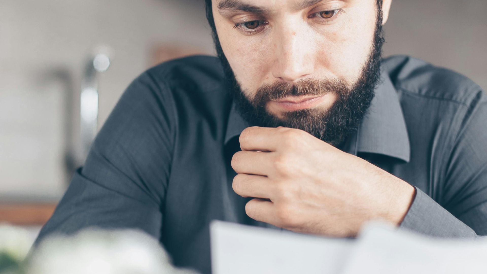 A bearded man in a gray shirt intently reads documents while seated indoors, appearing concerned.