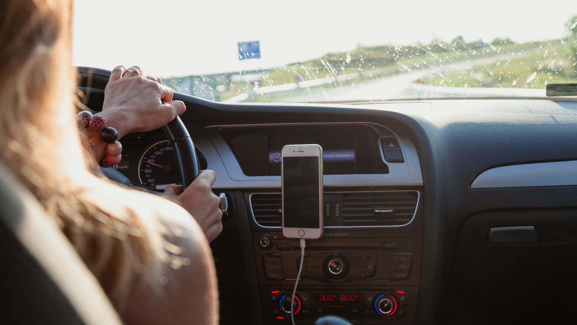 View from behind a woman driving a car on a sunny day with a phone mounted on the dashboard.