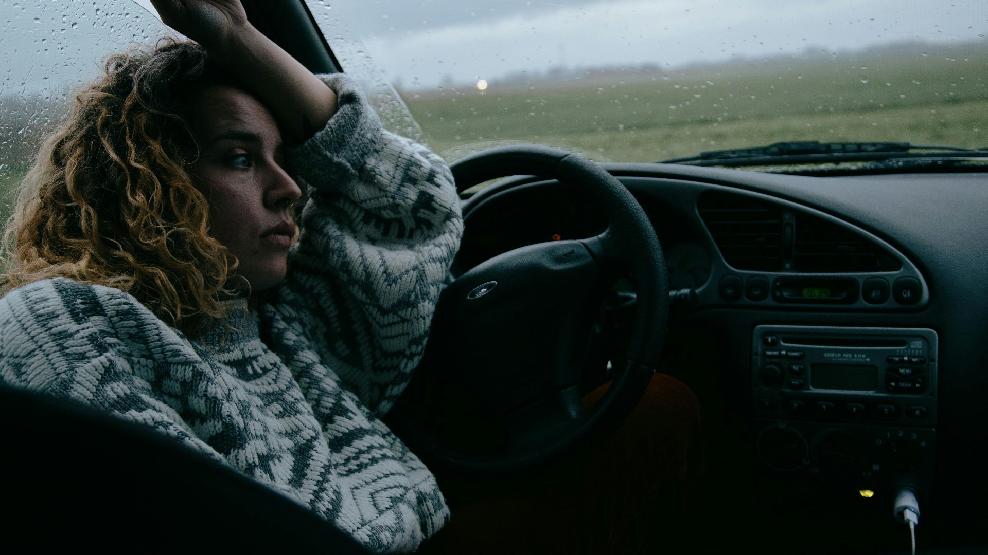 A woman sits in a parked car, gazing out of the window on a rainy day.