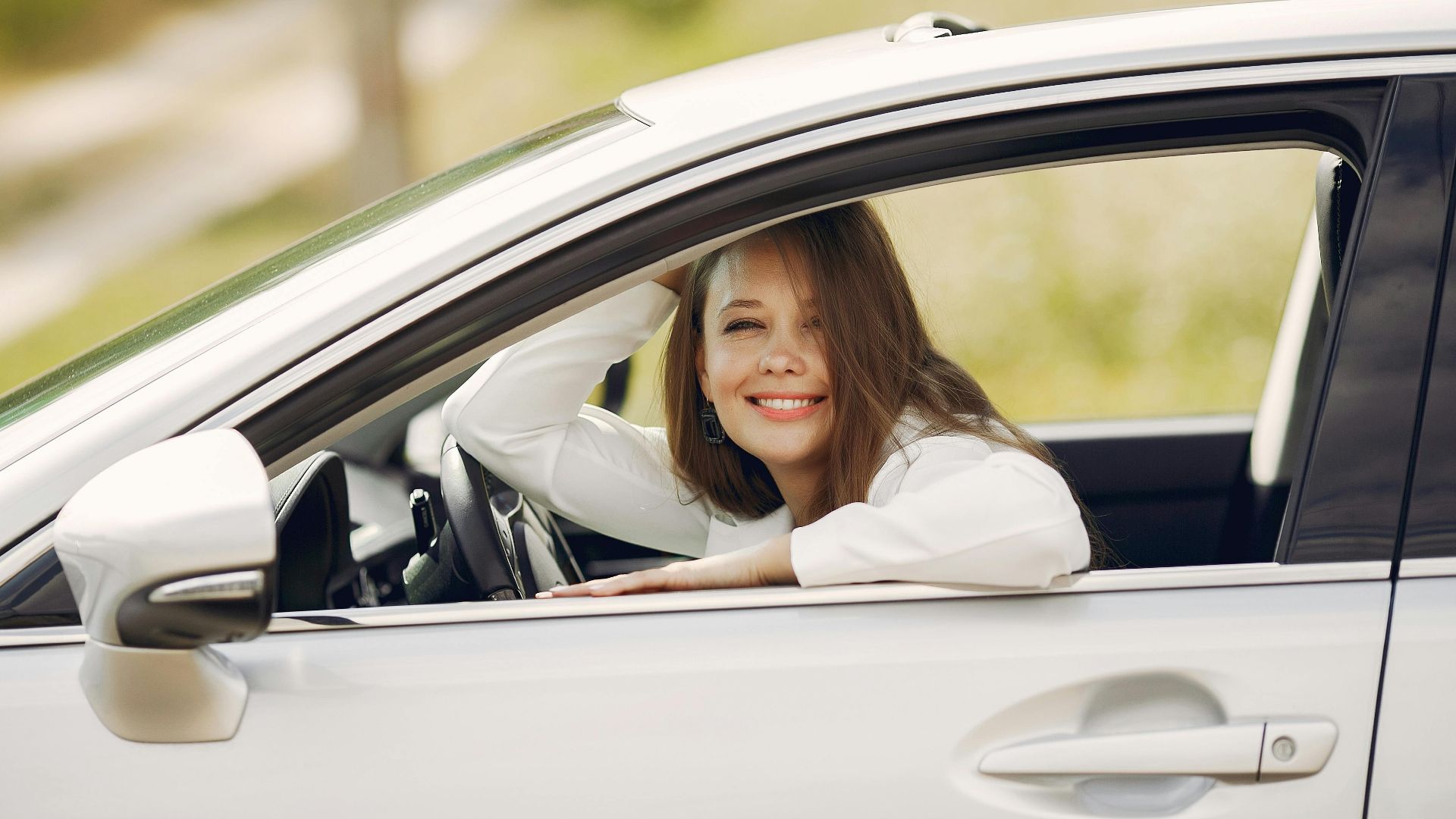 Cheerful female driver in white shirt sitting inside modern automobile and looking at camera through opened side window with smile during car trip in summer day