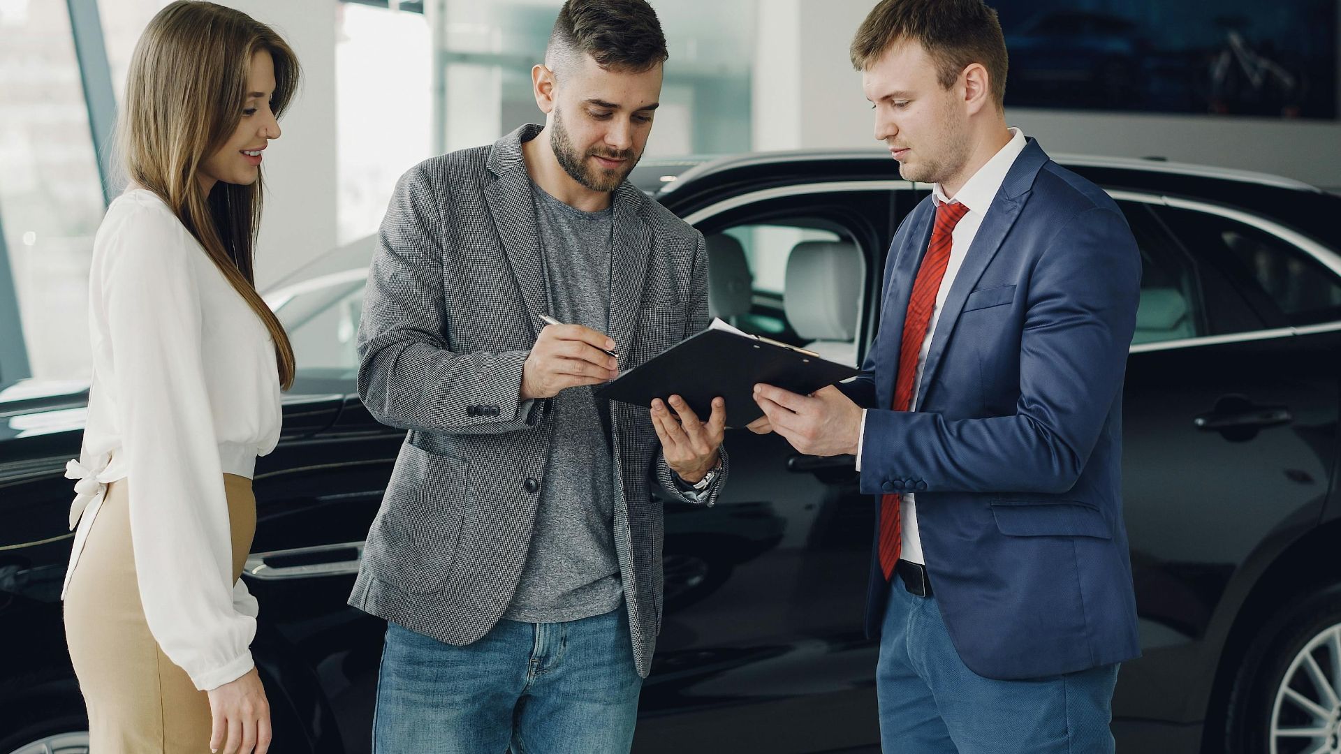 Three adults discussing documents at a car dealership beside a black car.