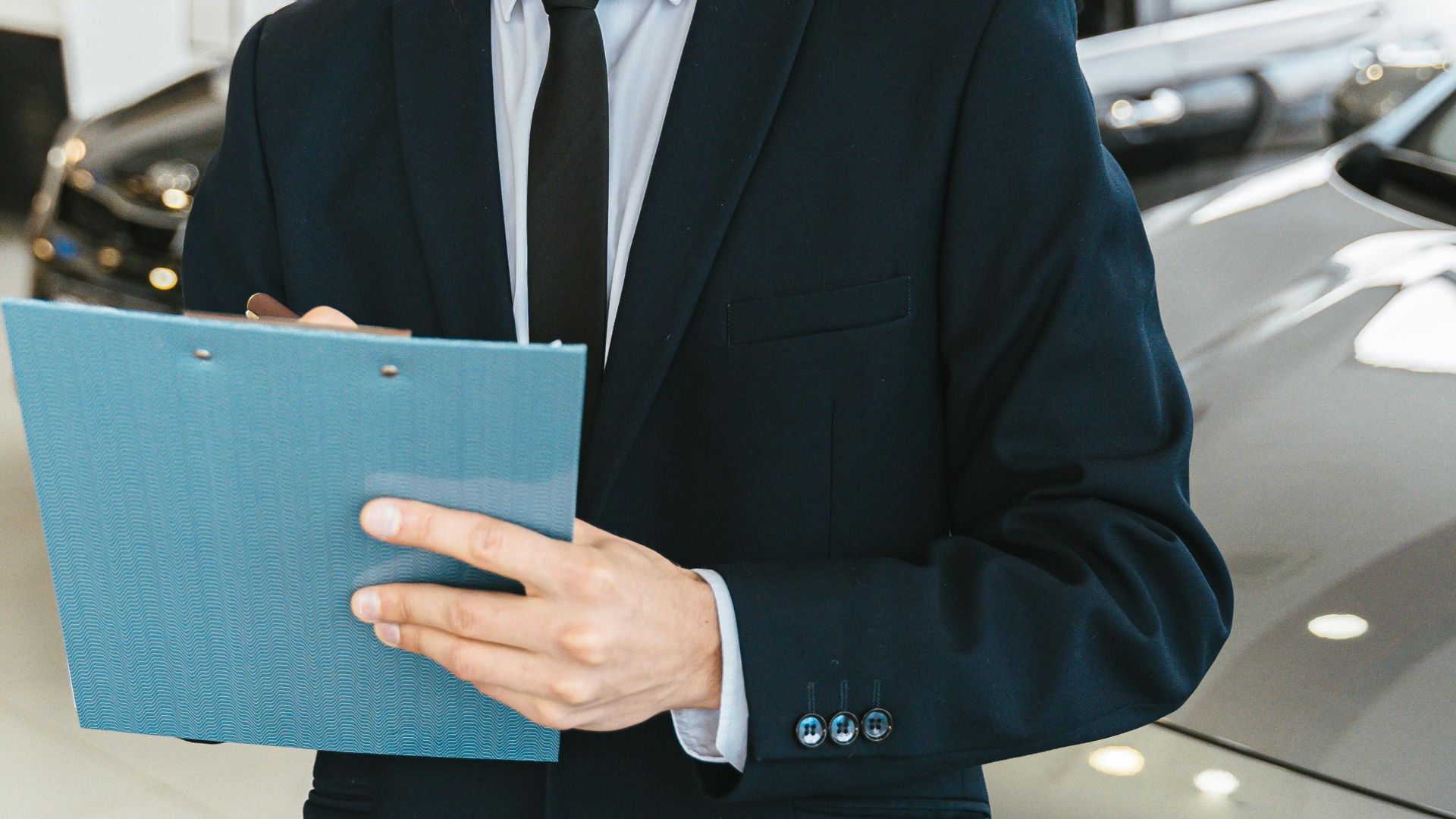 Confident car salesman in a suit with clipboard in a modern dealership showroom.