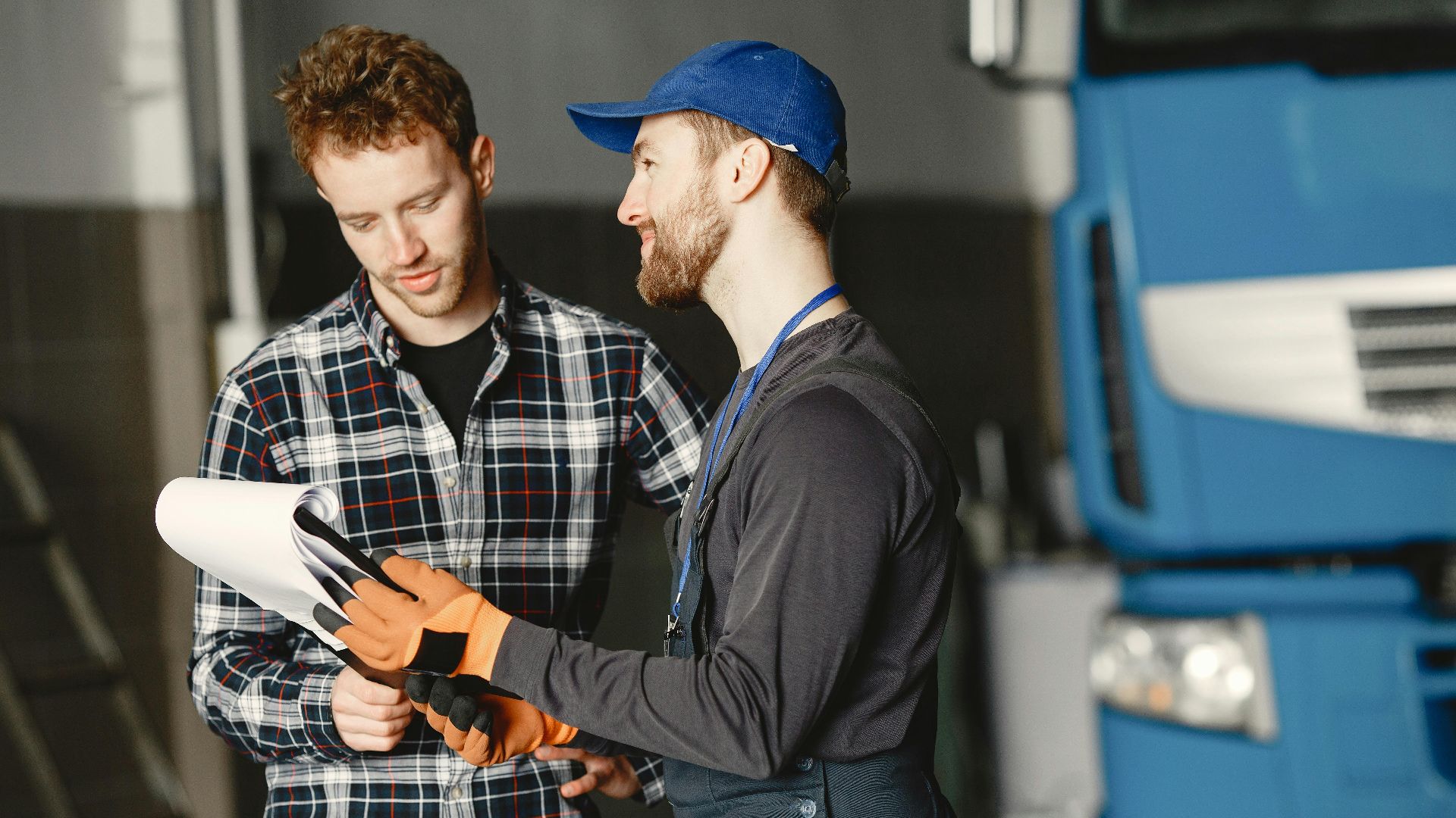 Mechanic in uniform consults with customer in garage setting beside a blue truck.
