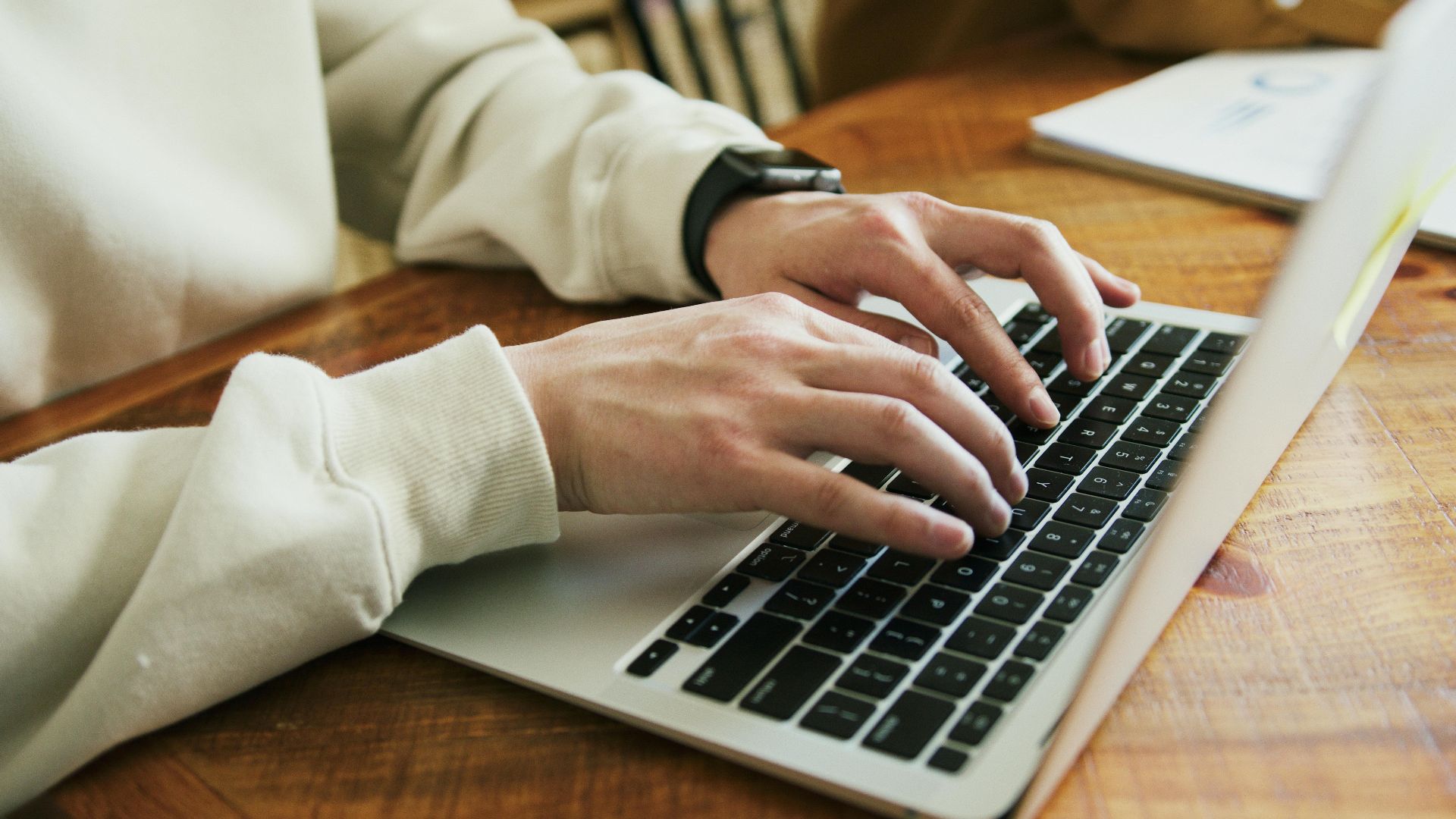 Hands typing on a laptop keyboard on a wooden desk, close-up view.