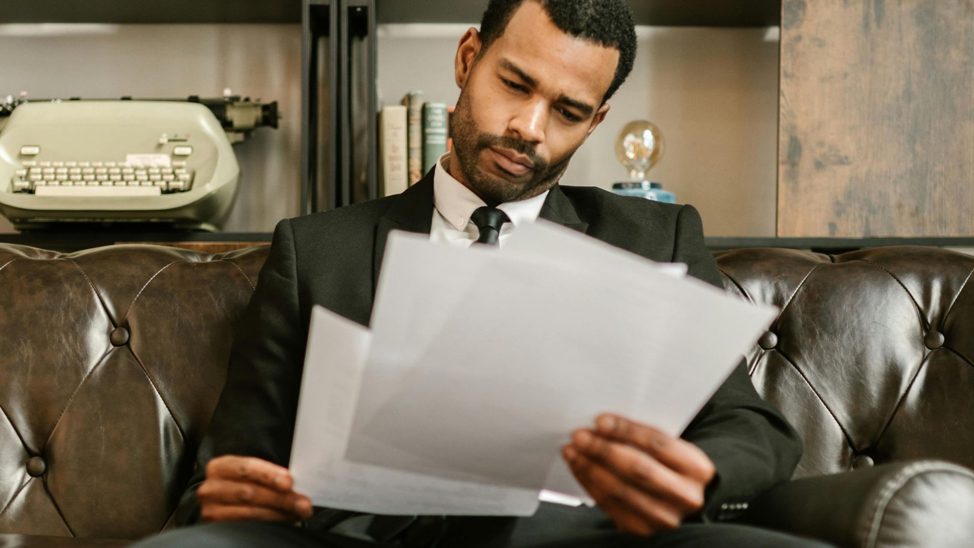Adult man in a suit reading paperwork on a leather couch in a stylish office setting.