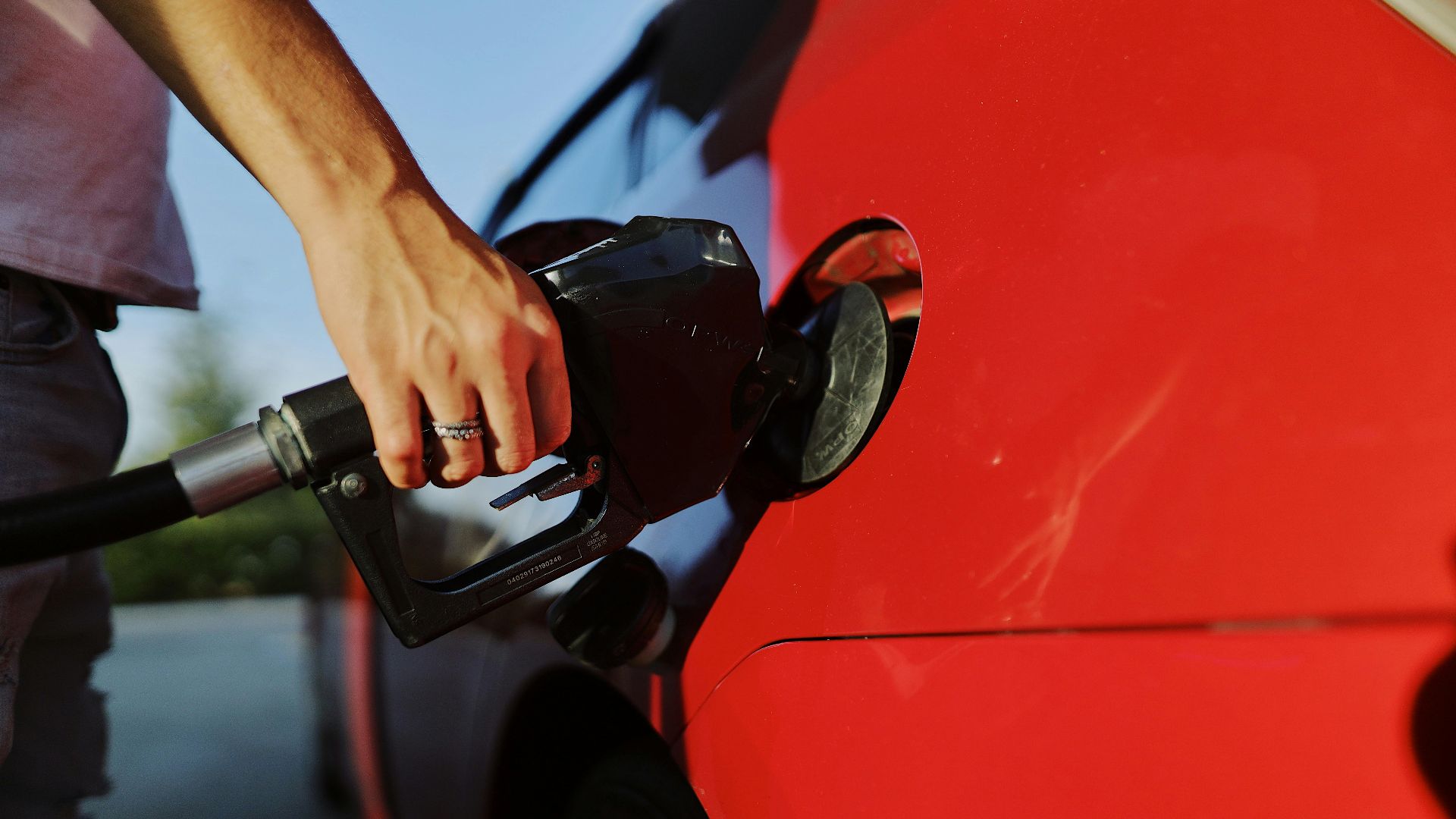 Close-up of a person refueling a red car at an outdoor gas station during the day.