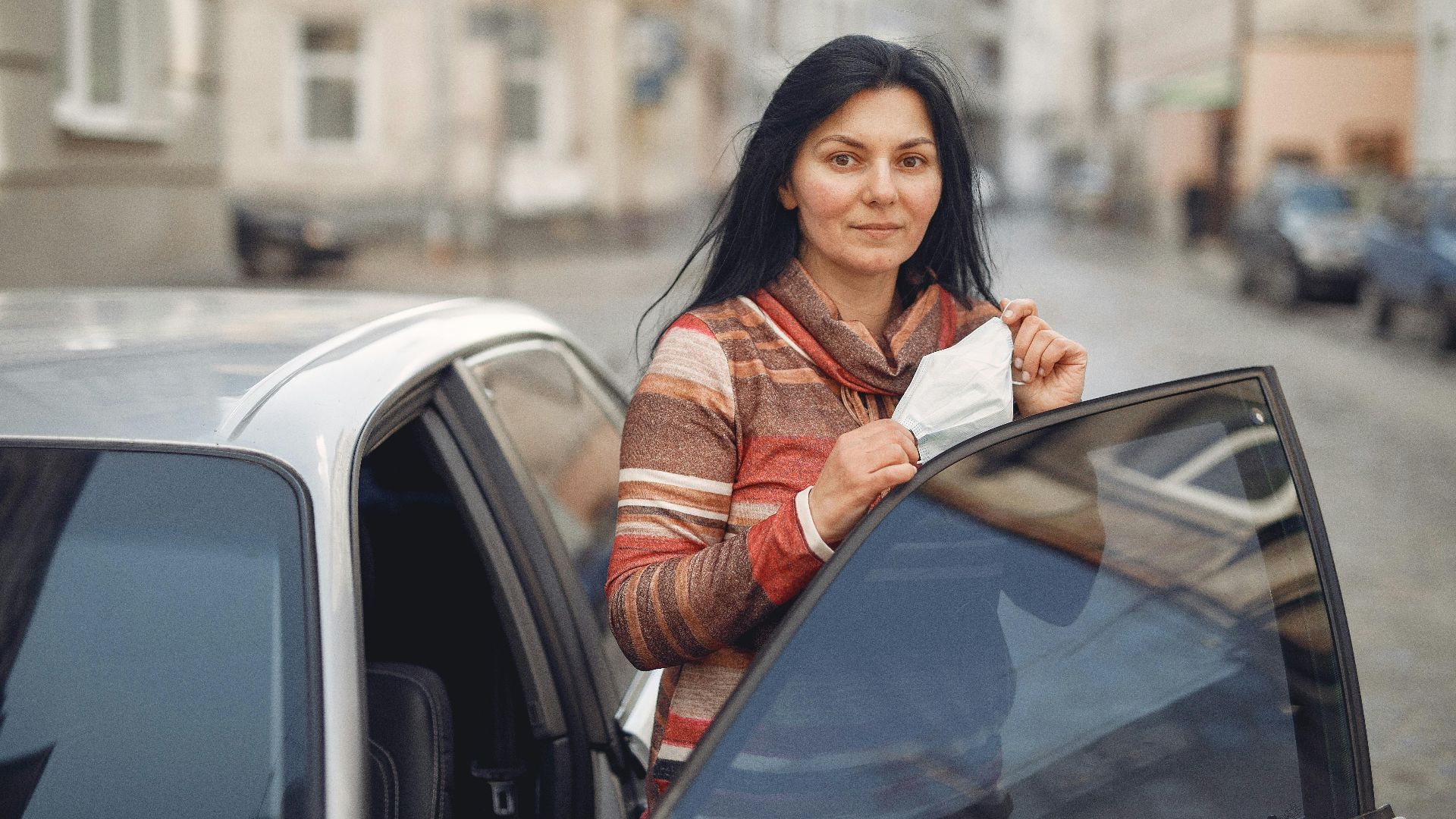A woman stands beside an open car door in a city street, holding a protective face mask.