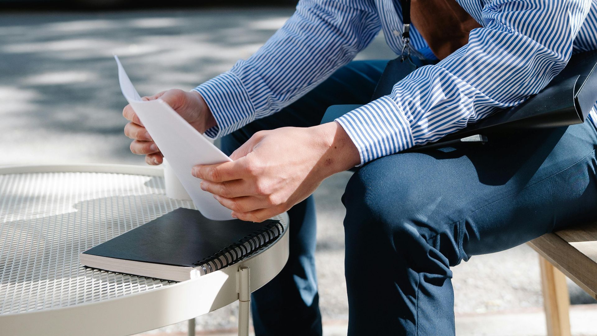 Asian man in striped shirt sitting outdoors reviewing documents at a table.