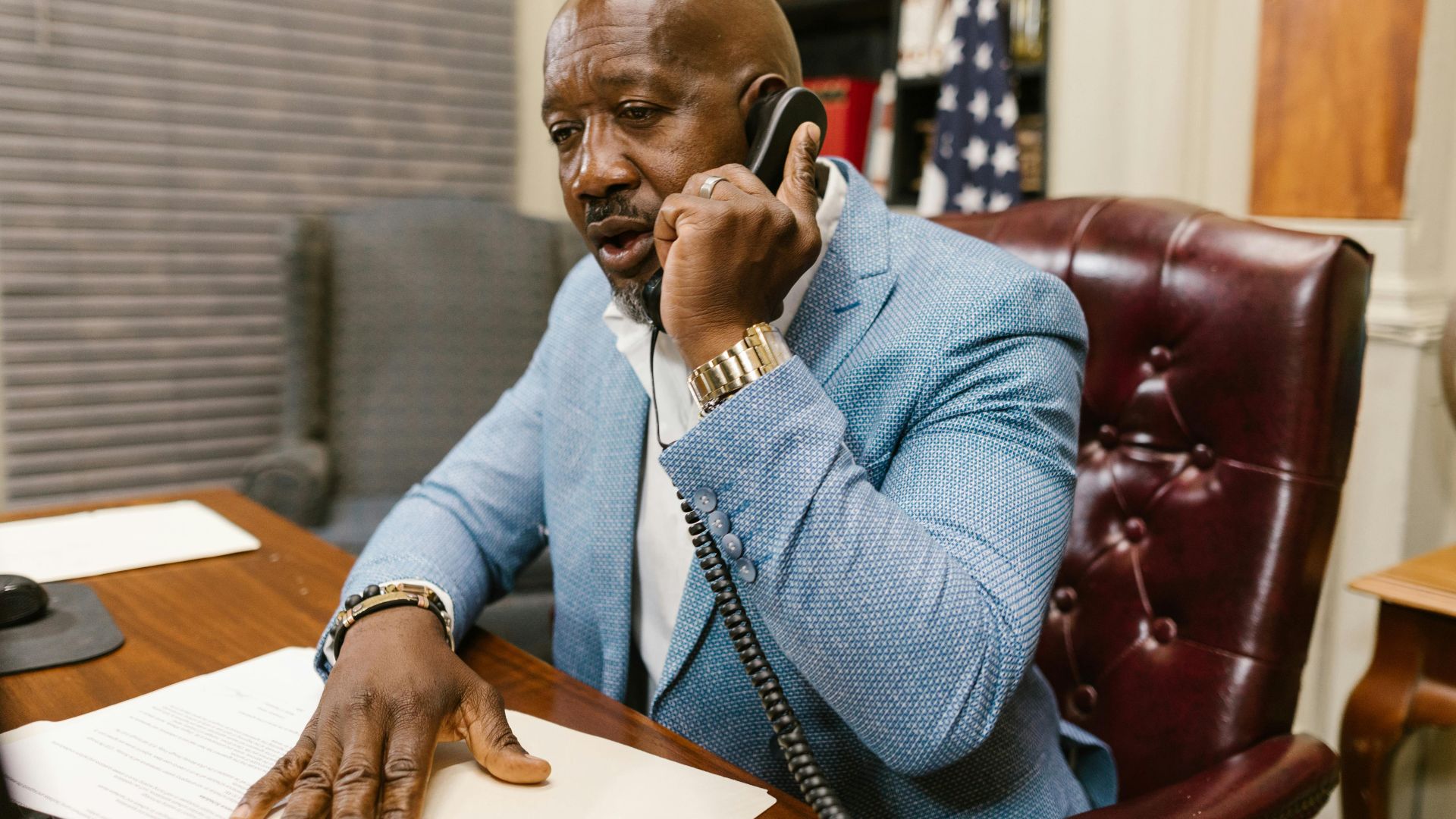 African American man in a blue suit speaking on the phone in a professional office setting.