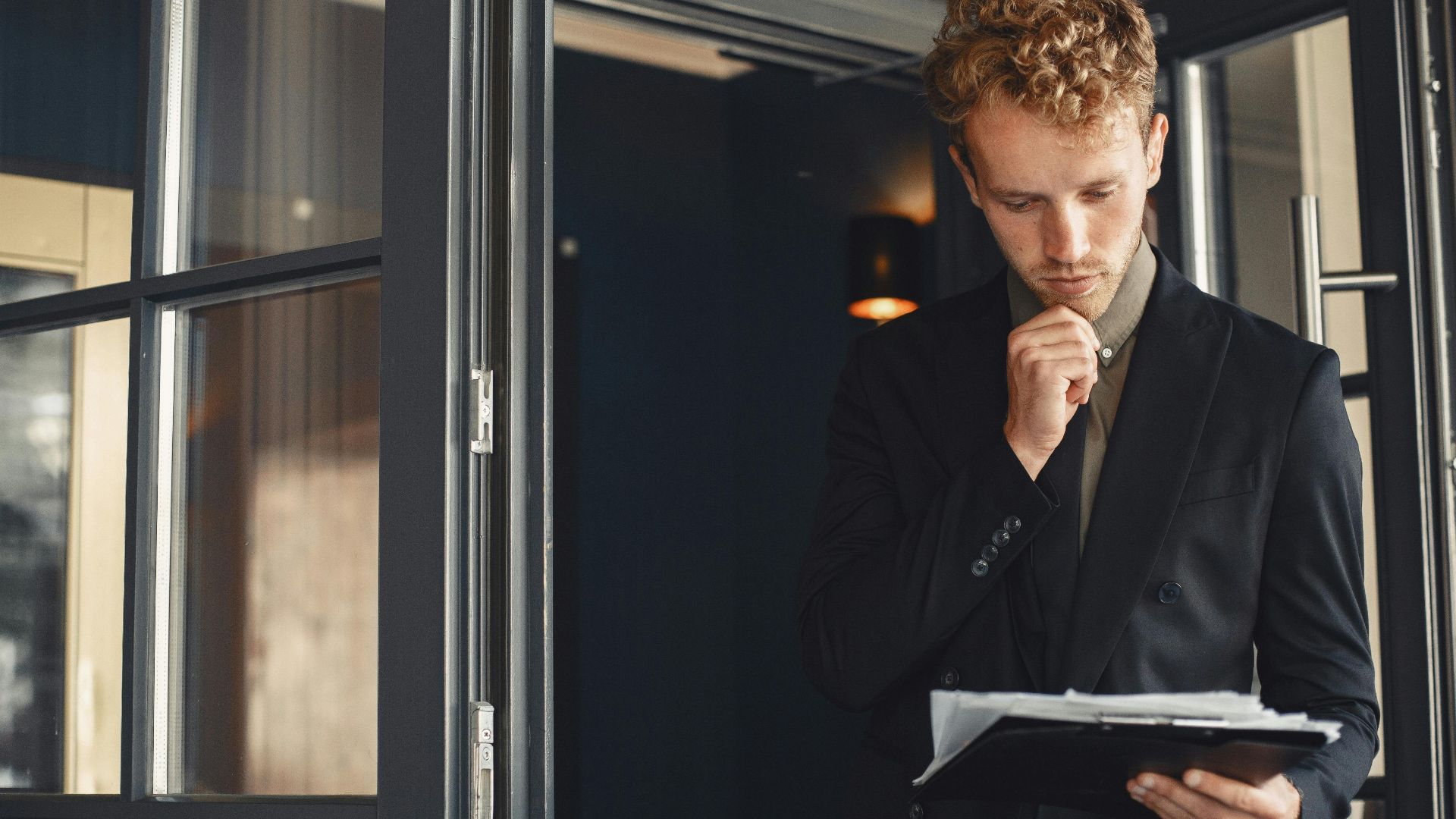A businessman in a suit thoughtfully reviews documents while standing by a door outdoors.
