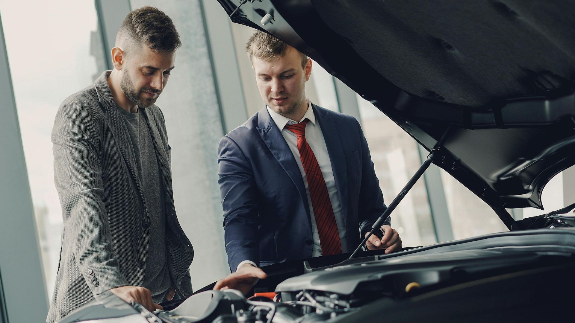 Two men inspecting a car's engine hood in a dealership showroom.