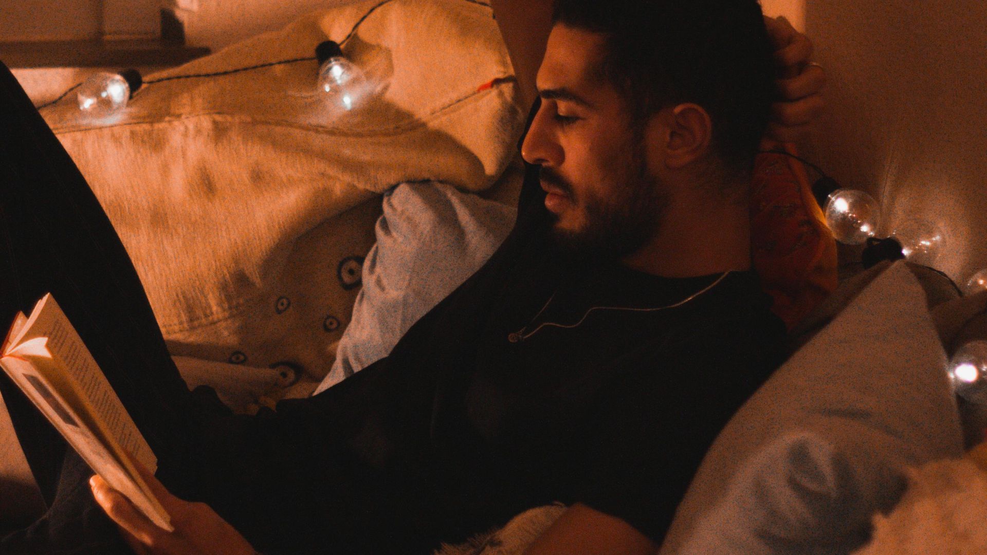 A man relaxes on a couch, reading a book under warm lights in a cozy room.