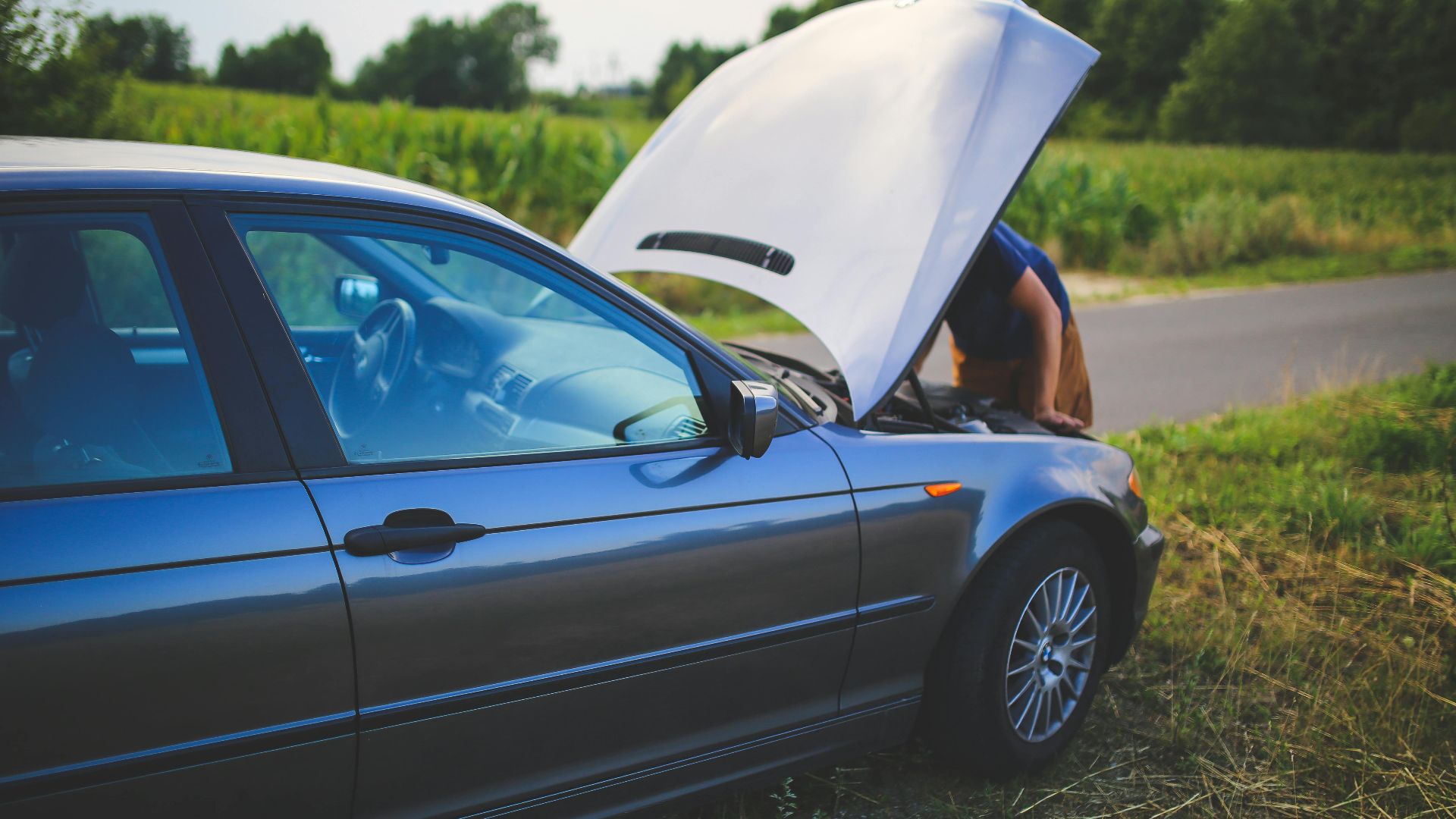 Man checking car engine with hood open by the side of a rural road.