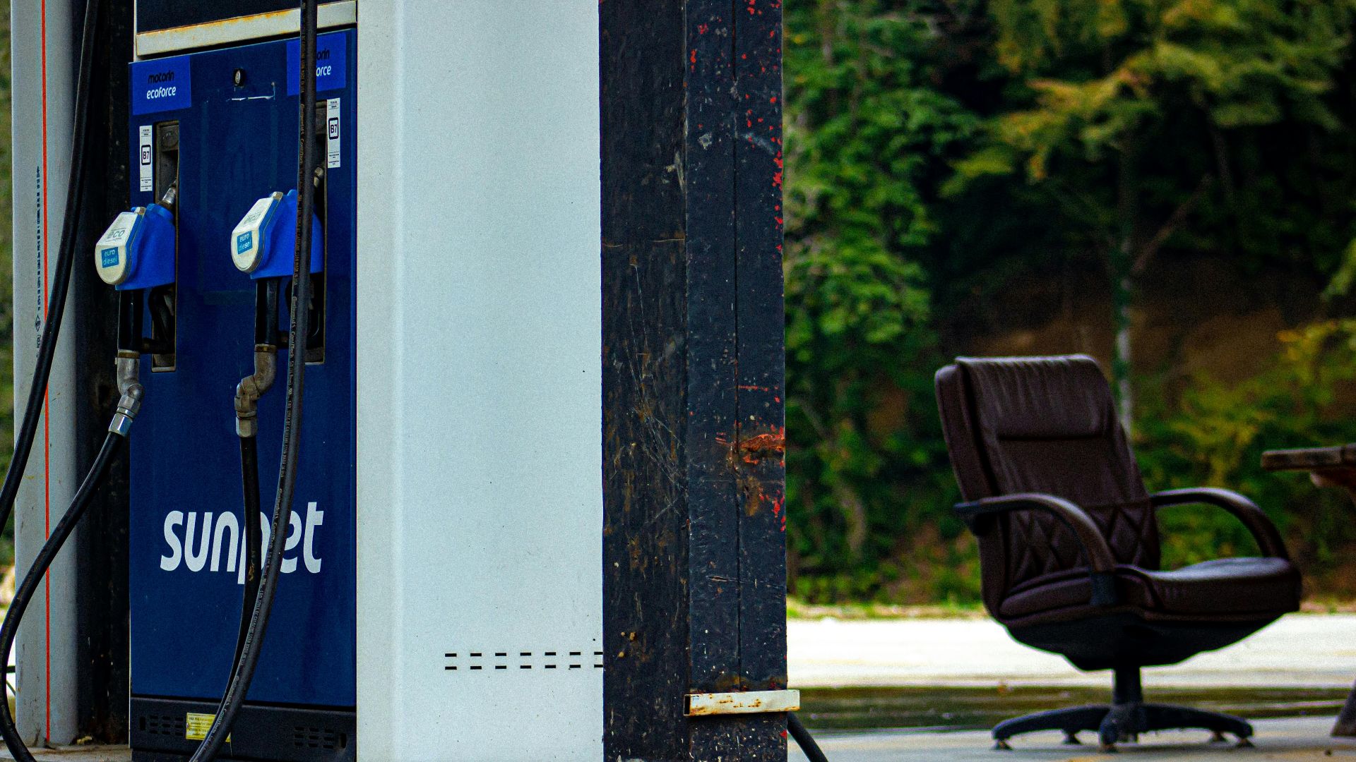 An outdoor gas station pump with a vintage look alongside an office chair, in a rustic setting.