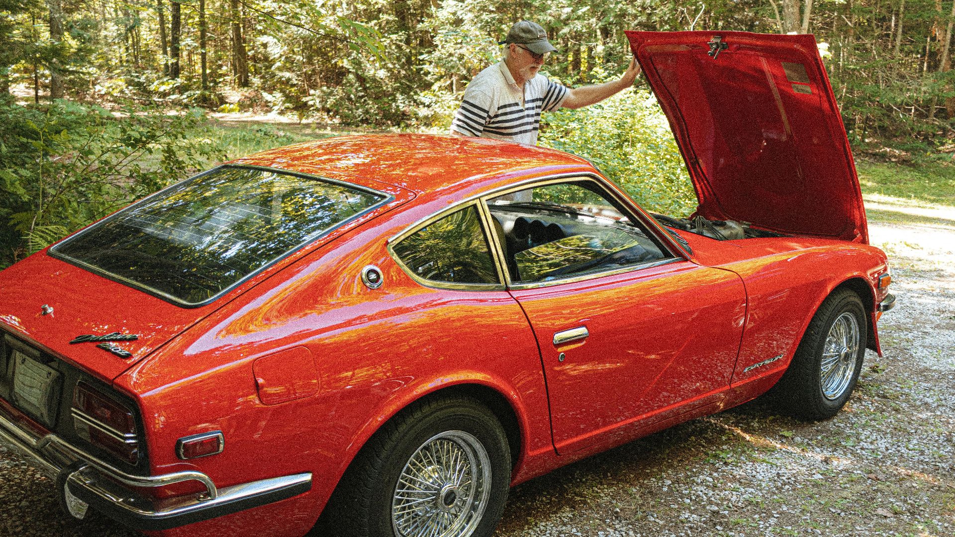 A senior man inspects the engine of a classic red Datsun 240Z parked outdoors.