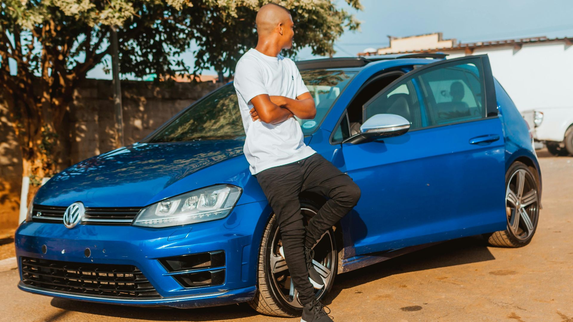 A young man in casual attire leans against a blue Volkswagen car parked outdoors on a sunny day.