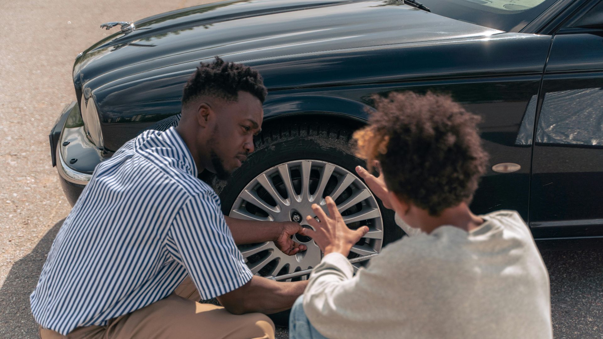 A father instructs his teenage son on how to change a car tire outdoors.