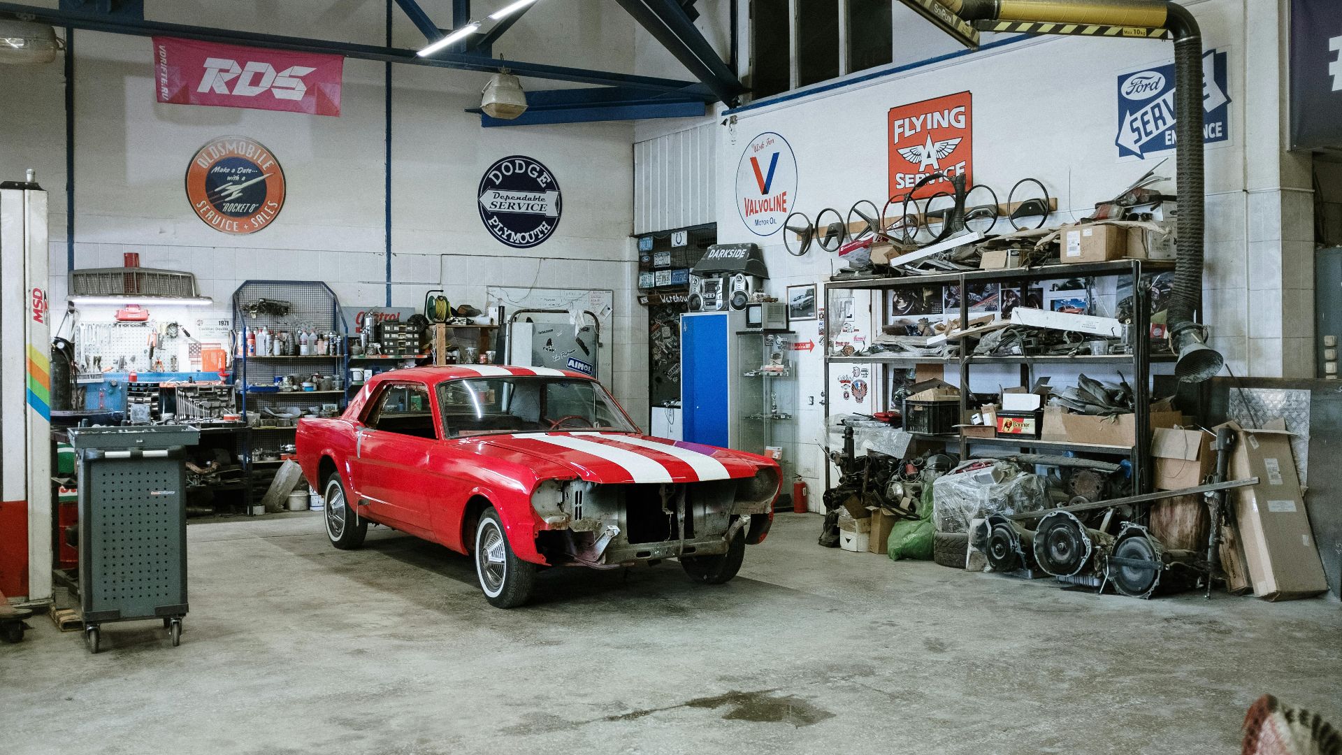 A vintage red car undergoing restoration in a well-equipped workshop with various tools and auto parts.