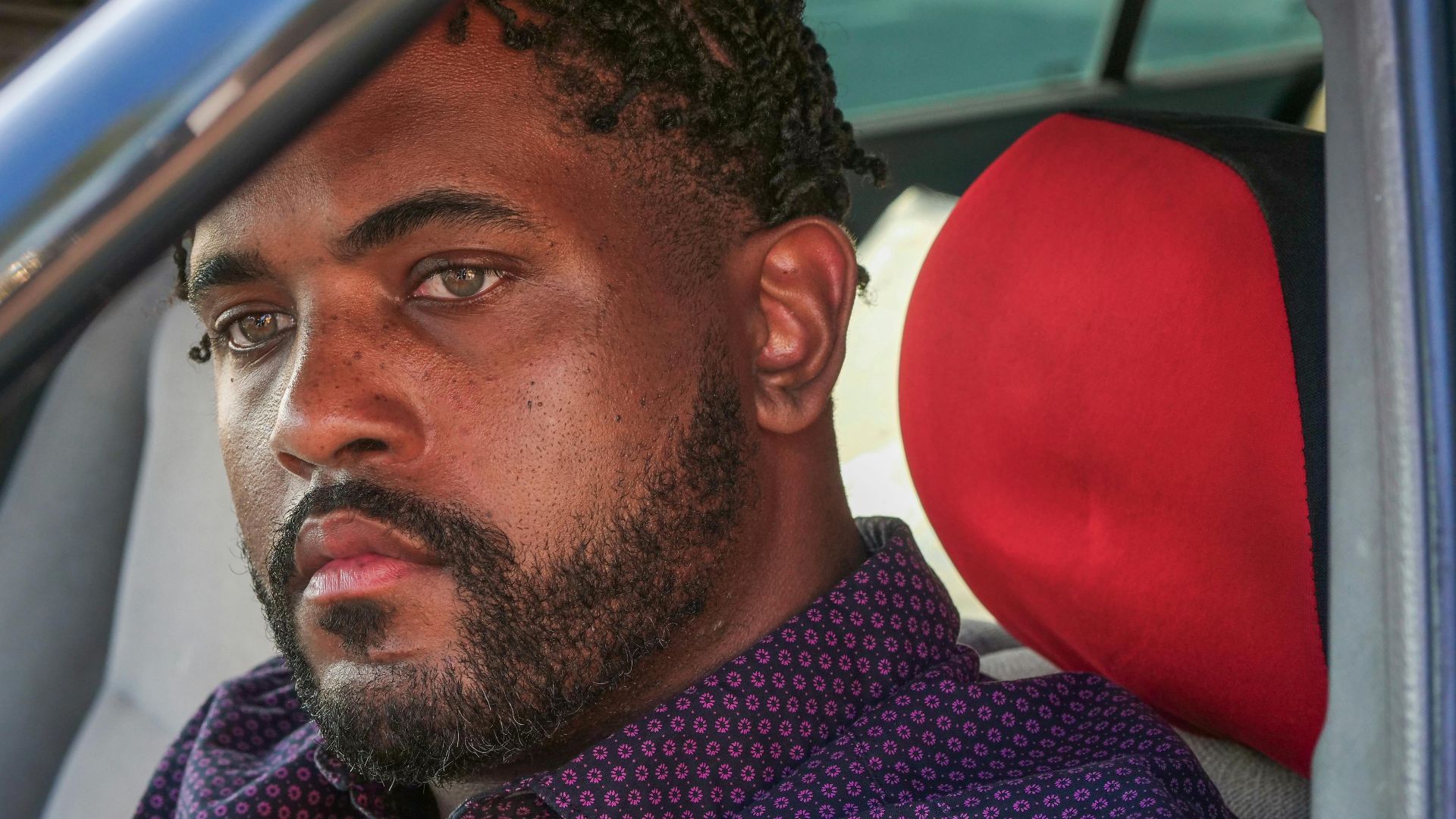 Close-up portrait of a man with facial hair sitting in a car staring intently.