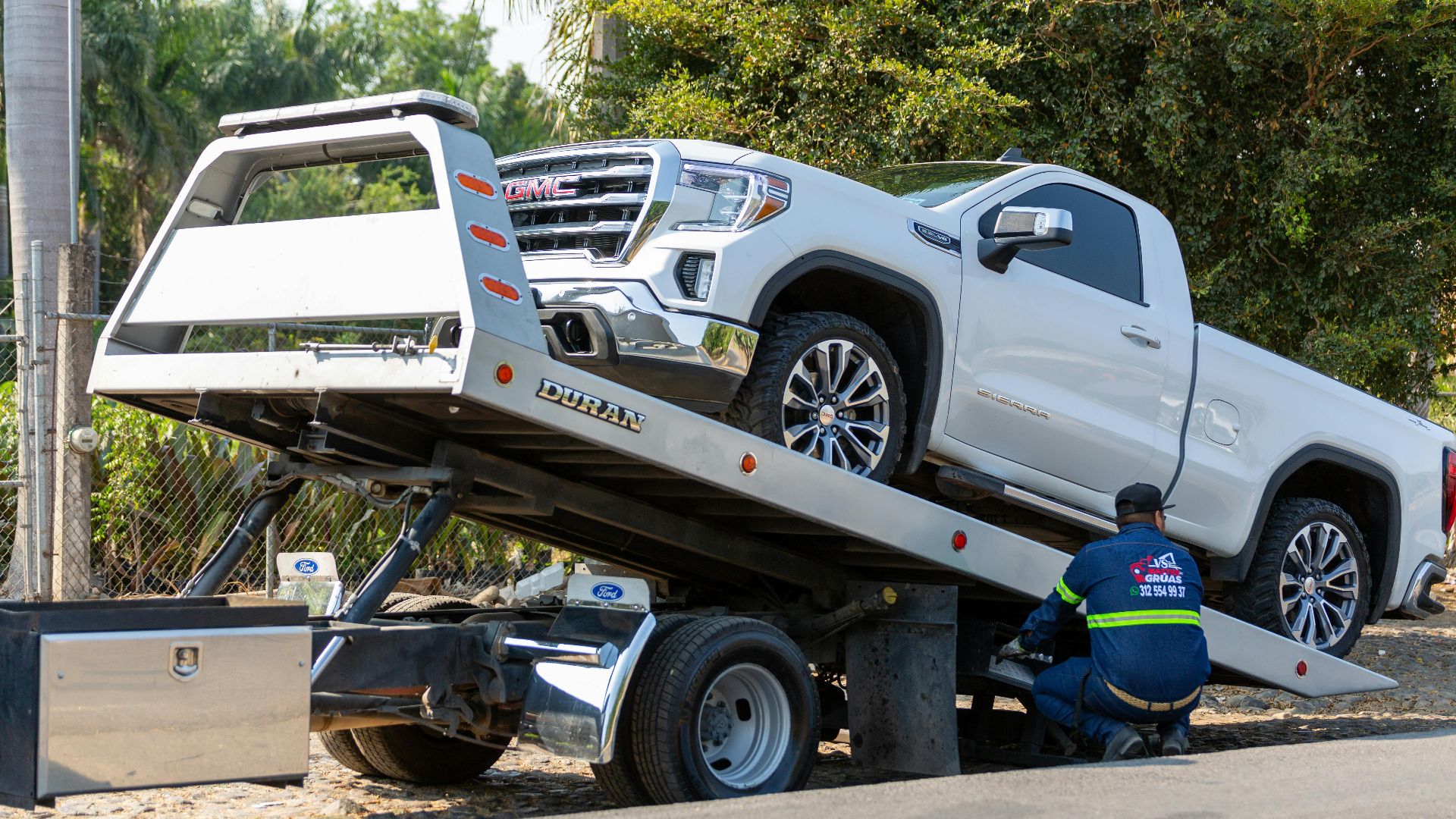Tow truck operator loading white GMC pickup truck on street in daytime.