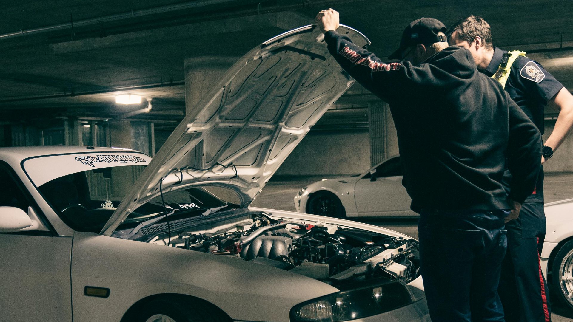 Two men examining a car engine in a dimly lit underground parking area.