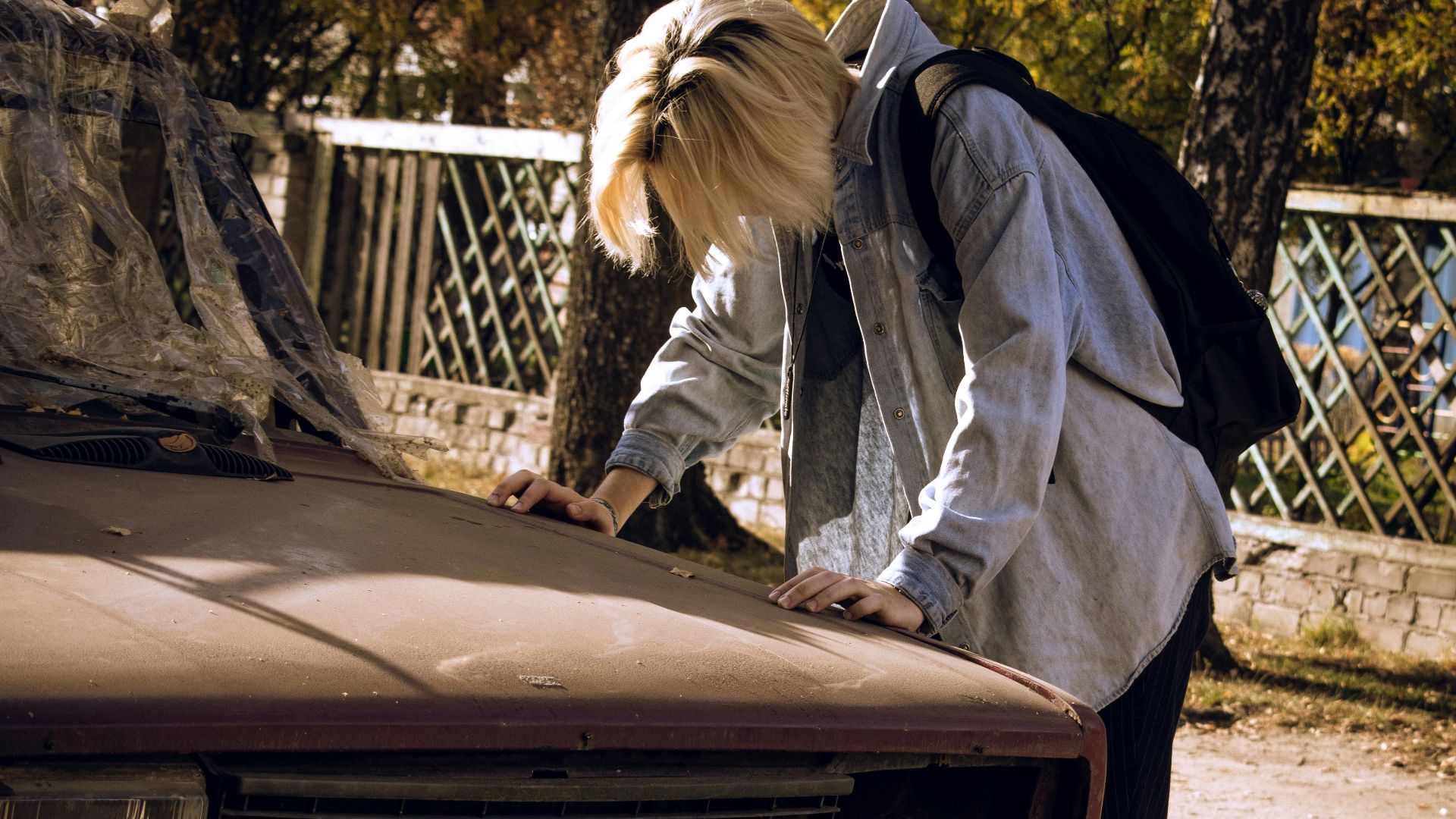 A young person leans on an abandoned car, reflecting stress and despair. Outdoors in natural daylight.