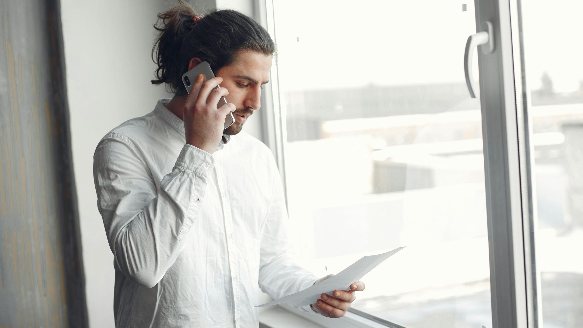 Young man in white shirt, on phone call holding a document, standing by a large window.