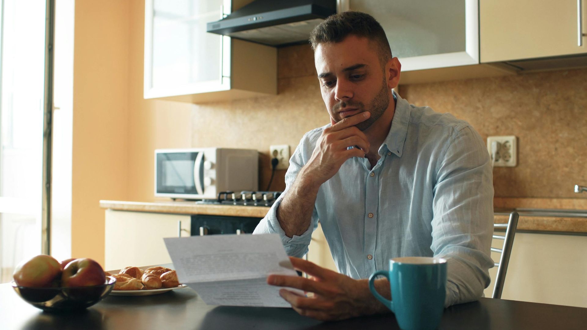 Thoughtful man reads a letter in a cozy kitchen setting with sunlight.