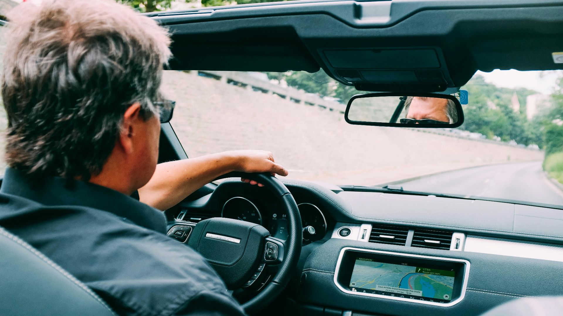 A man driving a convertible car on a scenic highway with a focus on the dashboard.
