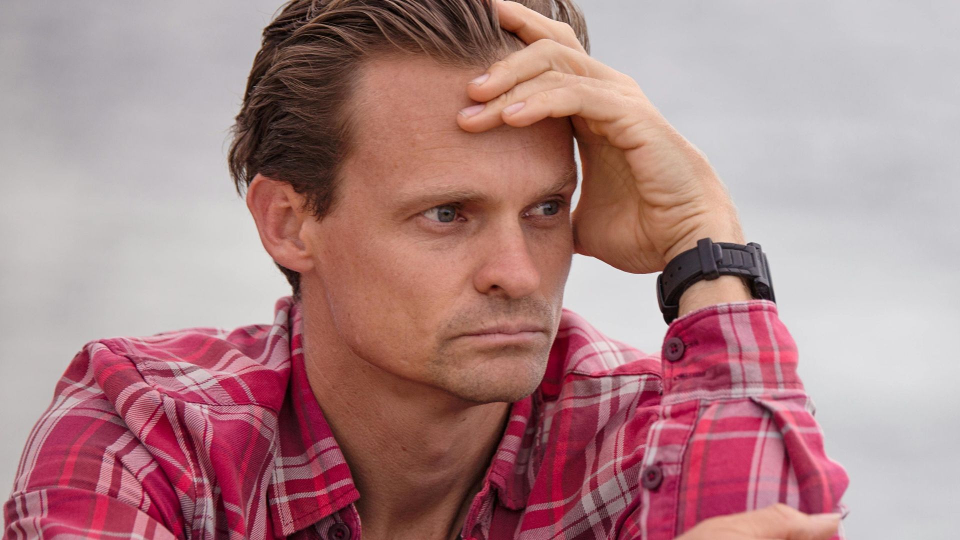 Adult male in plaid shirt sitting outdoors, deep in thought, with casual and relaxed demeanor.