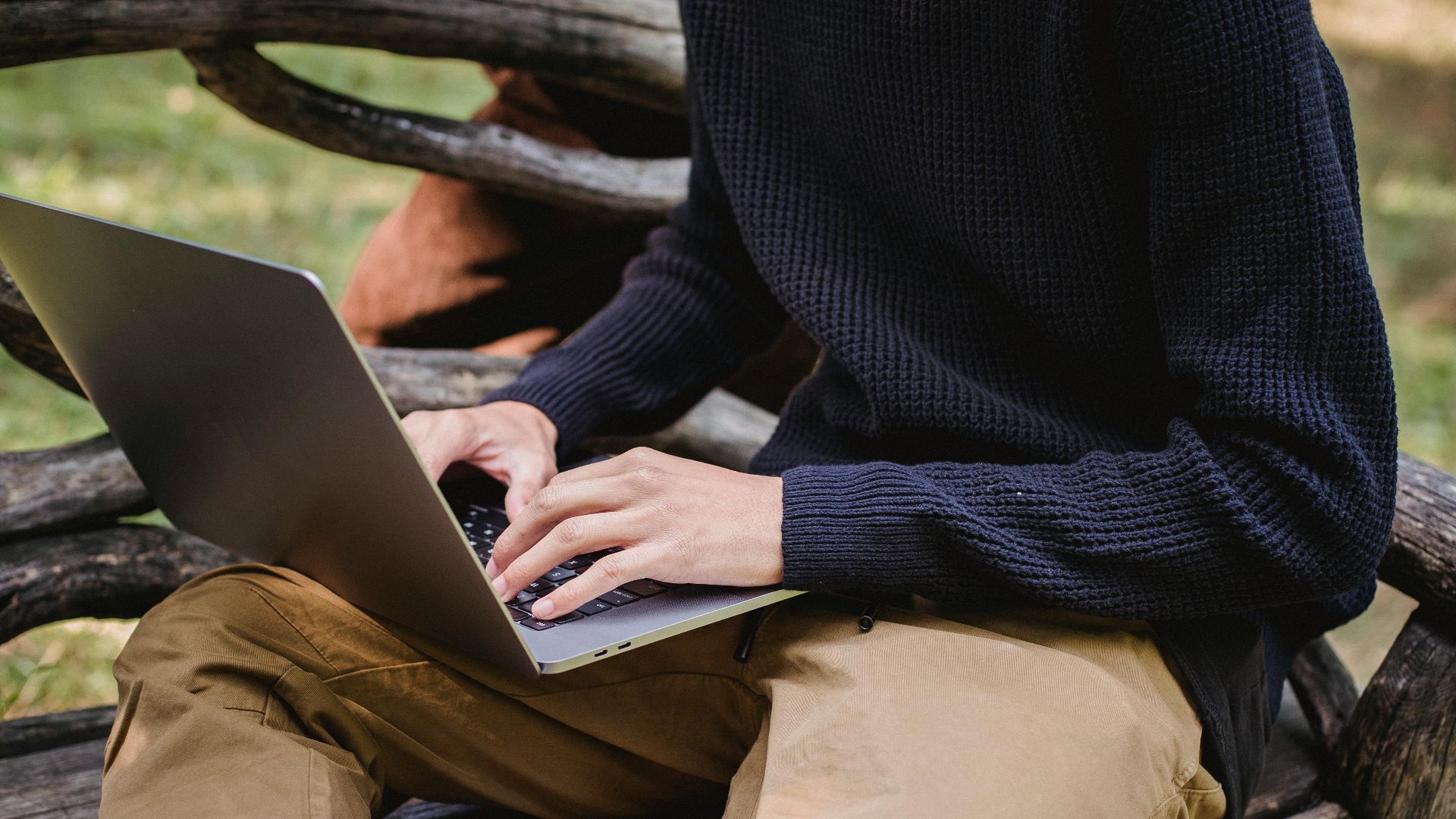 Crop anonymous male in casual clothes working on modern netbook while sitting on creative wooden bench in park