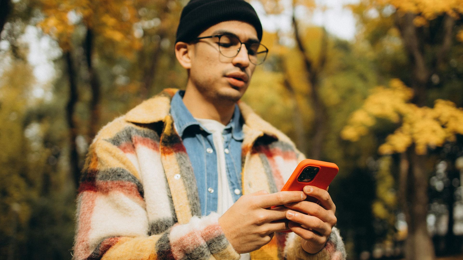 Young man in checkered jacket using smartphone outdoors in autumn park, focusing on mobile activity.