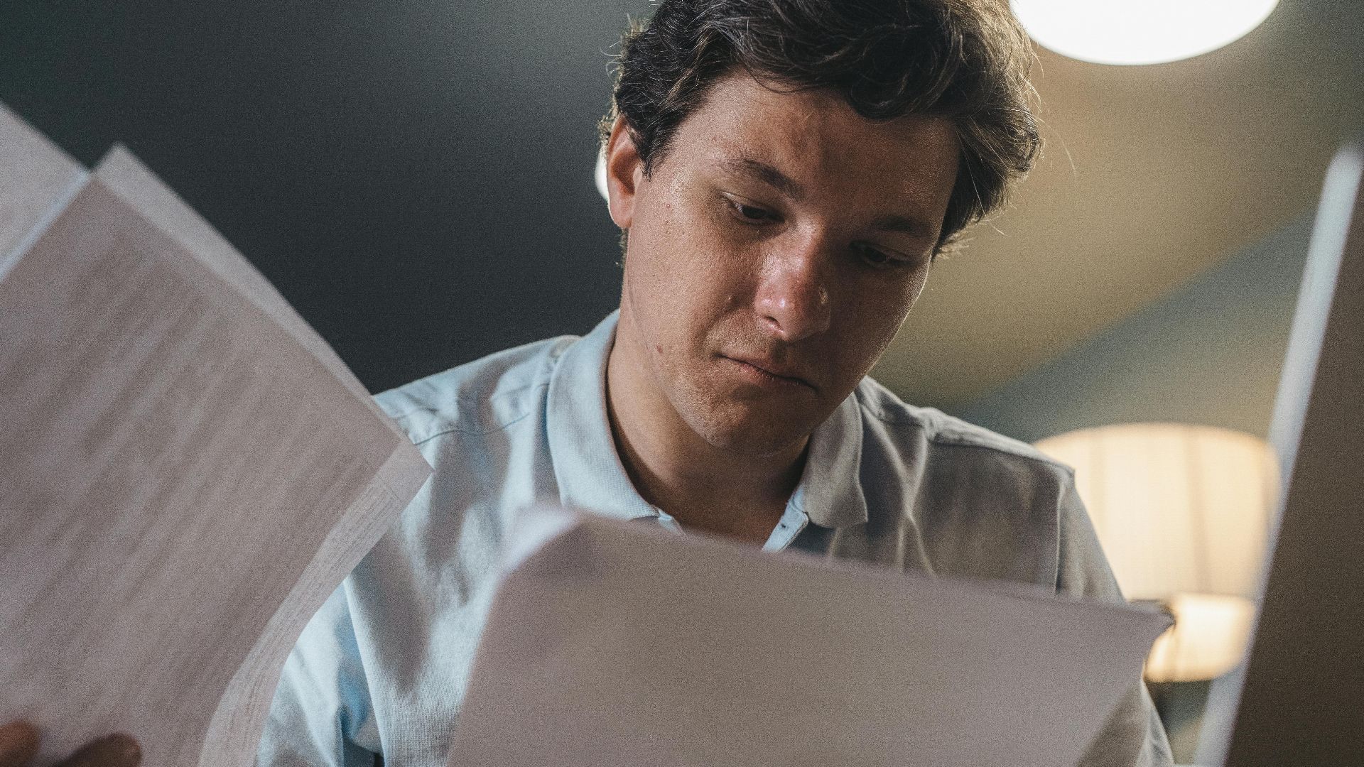 Serious man wearing a light blue shirt intently reading printed papers indoors under warm lighting.