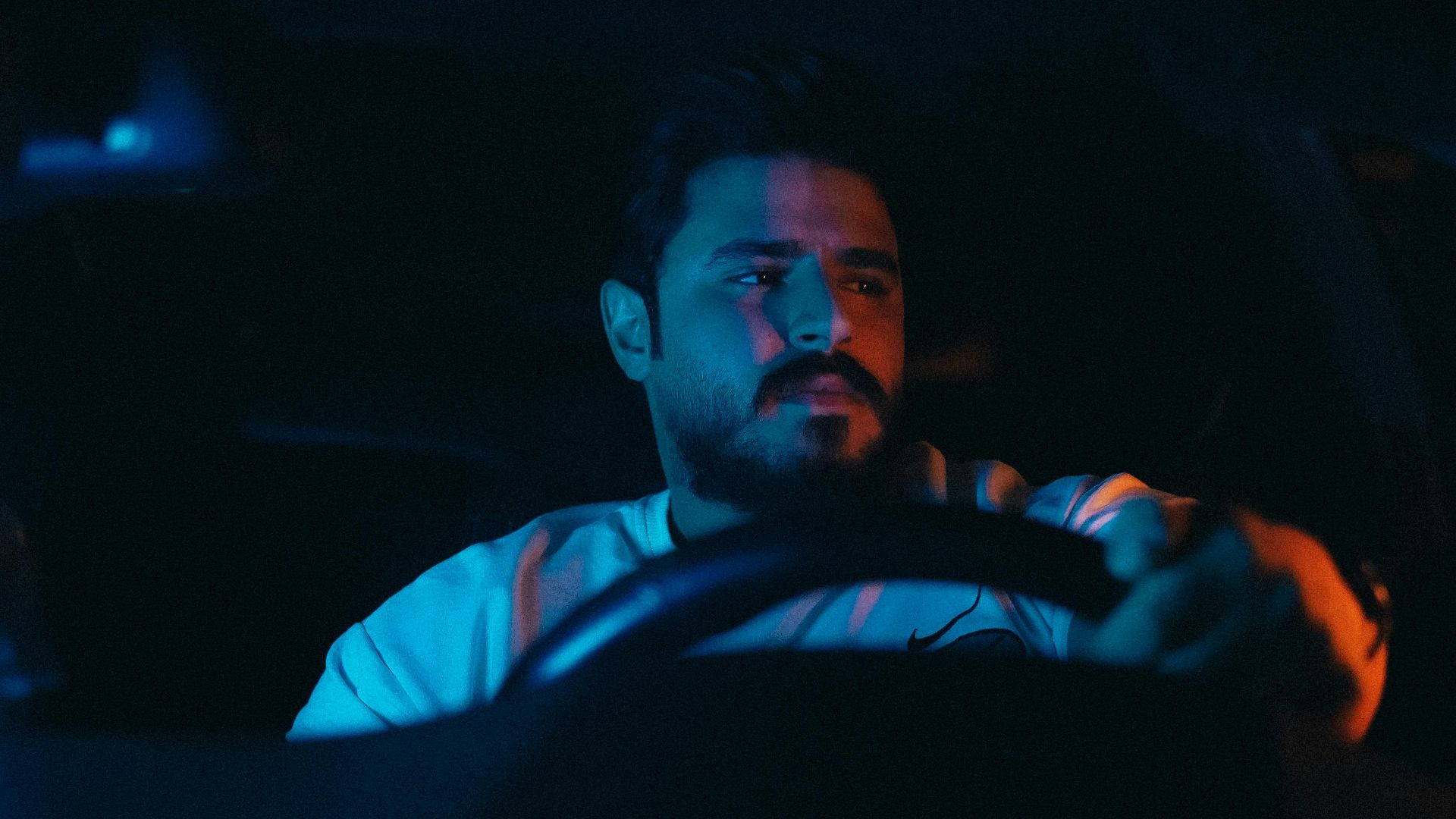 A focused man drives a car at night illuminated by moody lighting.